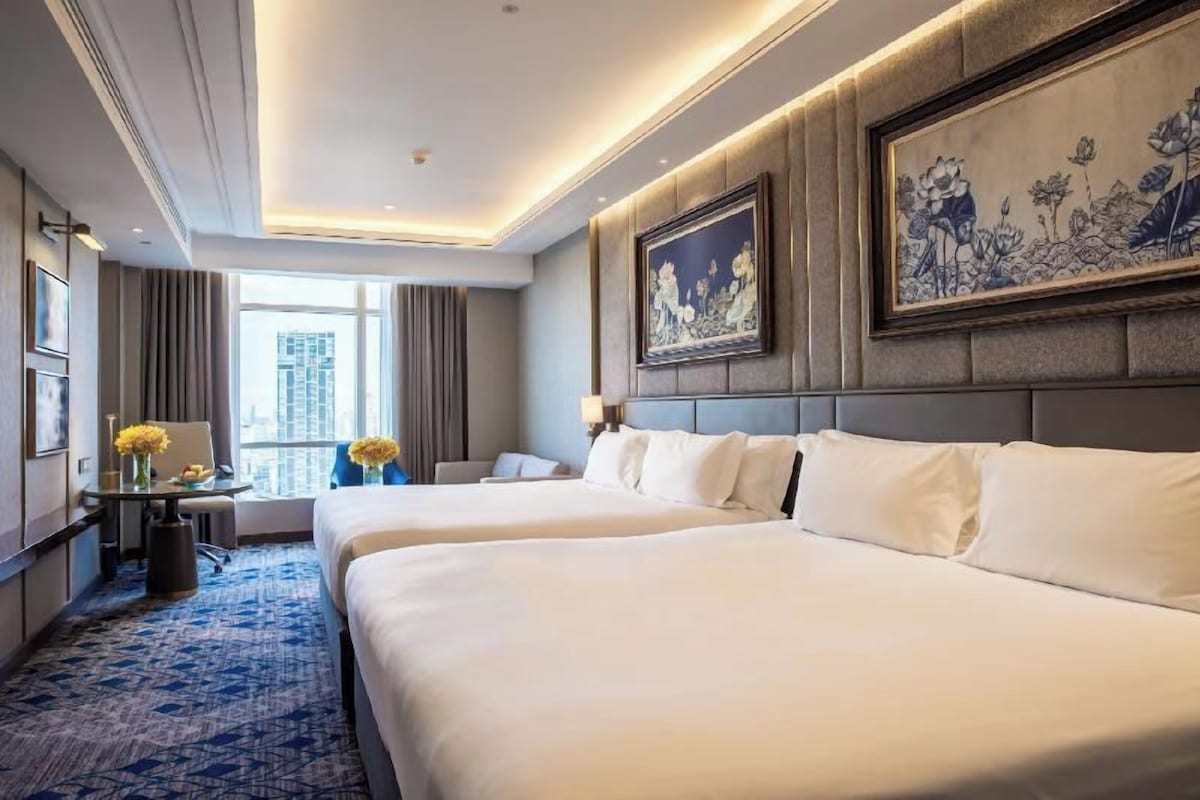 A hotel room with two beds, a large window, a painting on the wall, and a view of the city.