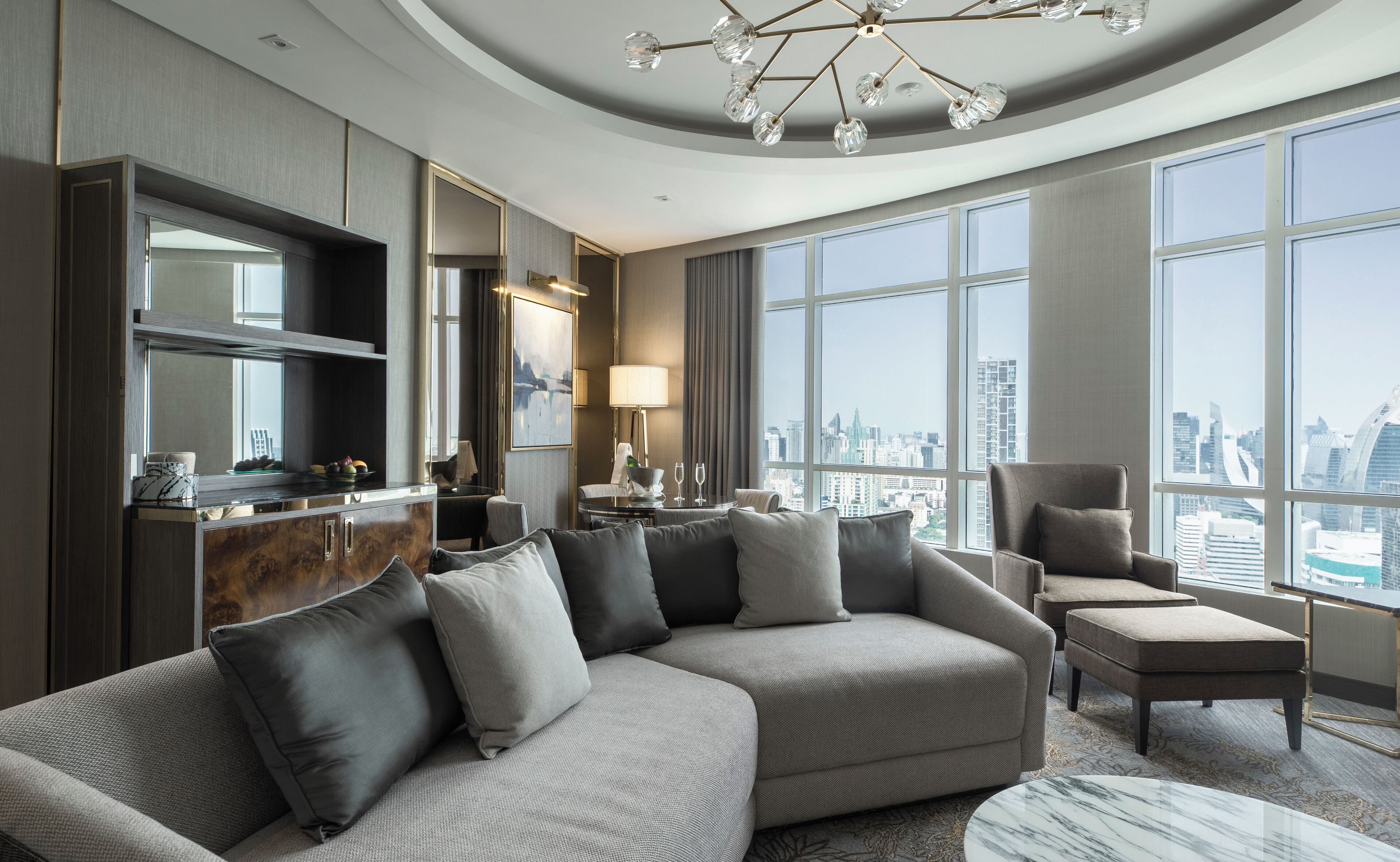 A modern hotel room with a sectional sofa, a dining area, a large window with a city view, and a chandelier.
