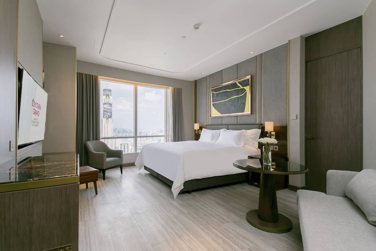 A modern hotel room with a large bed, a sitting area with a chair, a small table with a vase, and a large window offering a city view.