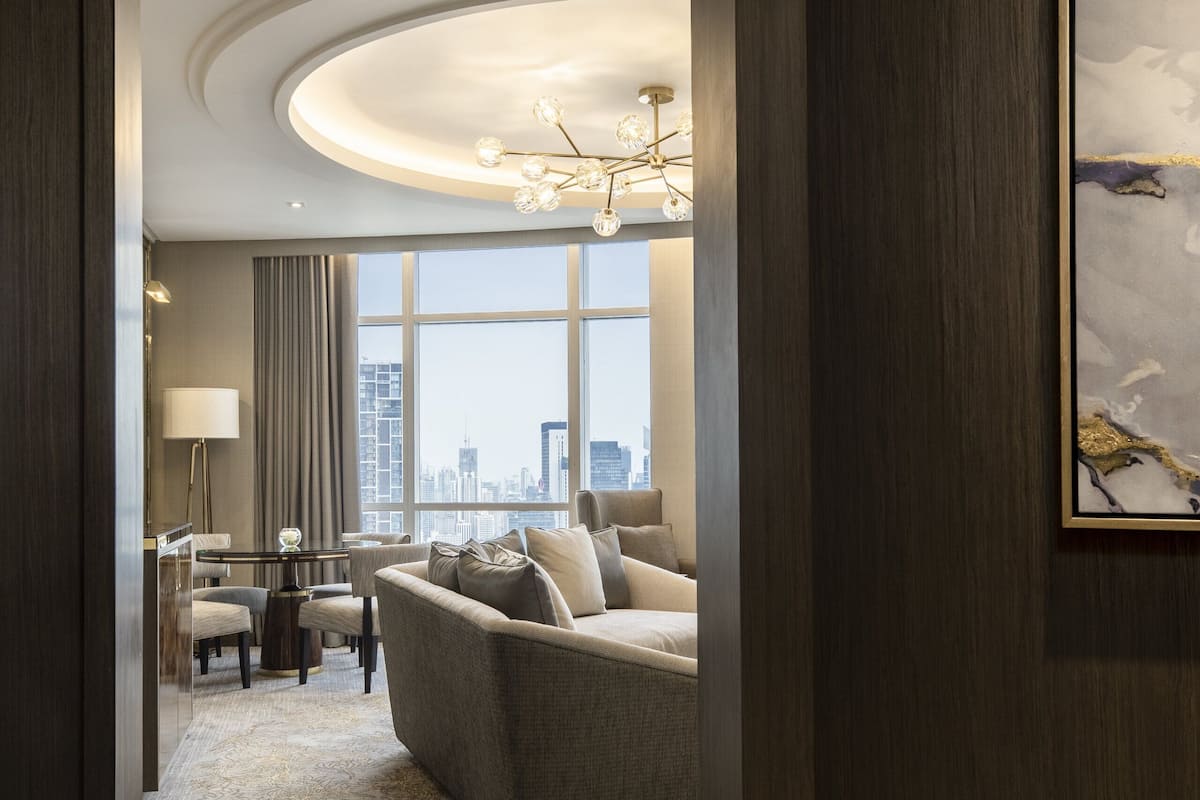 A hotel room with a city view, a sofa, a dining area, and a chandelier.