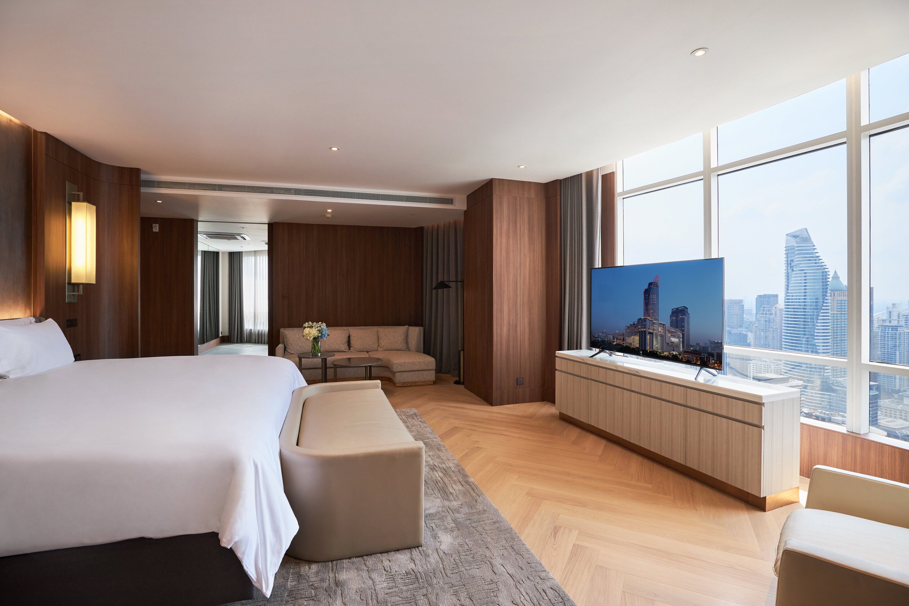A modern hotel room with a large bed, a sofa, a TV, and a city view.