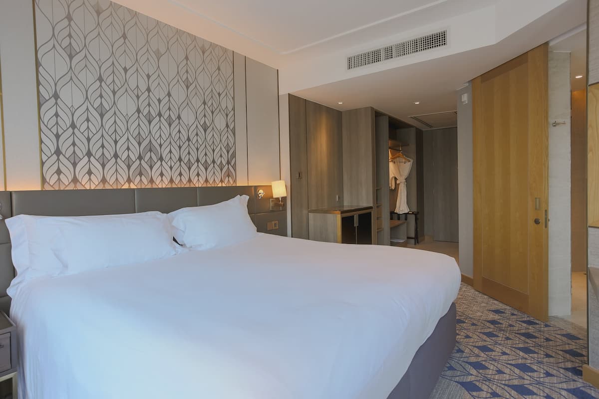A large bed with white linens, a headboard with a leaf pattern, and a bedside table with a lamp.