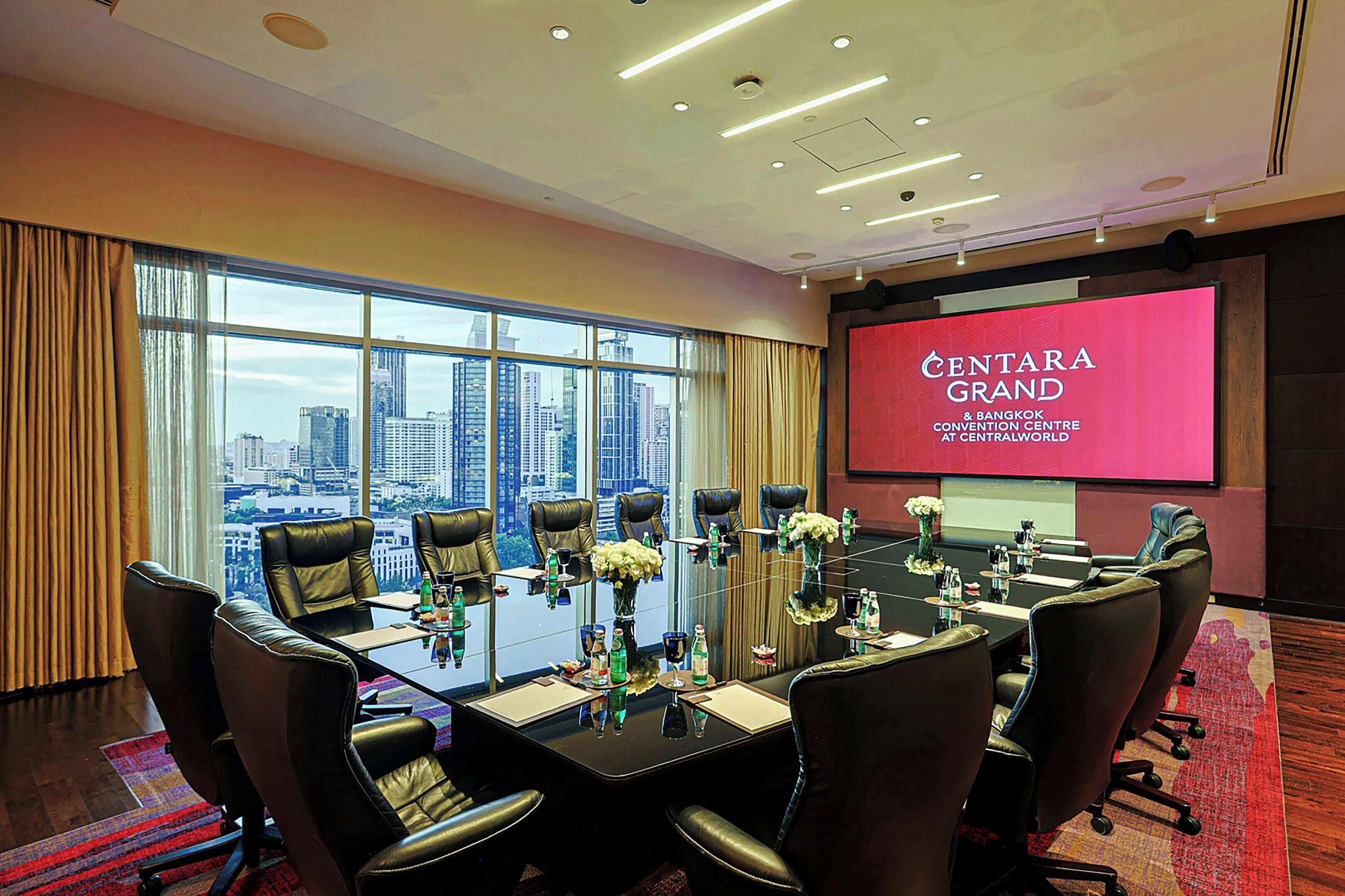 A conference room with a city view, a large screen displaying 'CENTARA GRAND & BANGKOK CONVENTION CENTRE AT CENTRALWORLD', and a table set for a meeting.