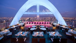 Un área de comedor en la azotea con mesas y sillas, una gran estructura en forma de arco blanco y una vista panorámica de la ciudad al fondo.