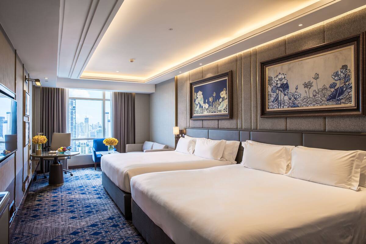 A hotel room with two beds, a desk, a chair, and a large window with a city view.