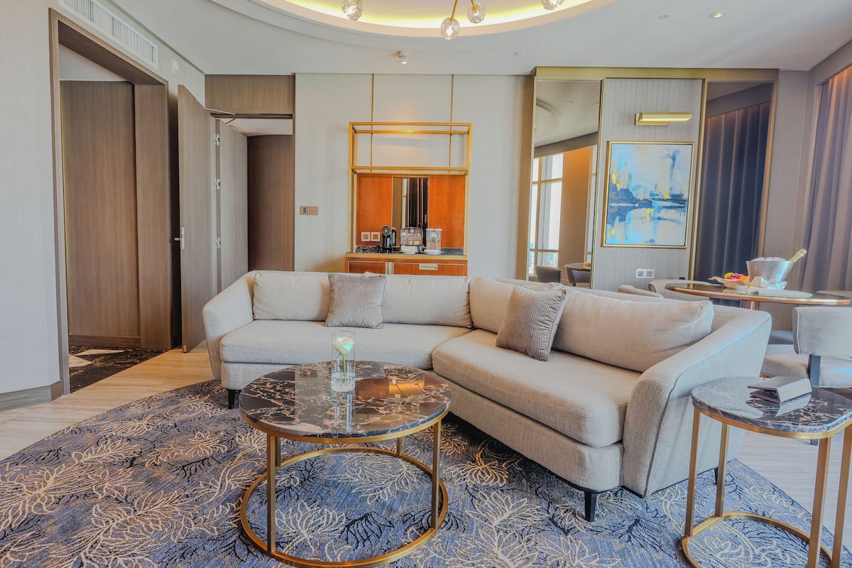 A modern hotel room with a large L-shaped sofa, a round coffee table, and a small side table. There is a dresser with a mirror and a painting on the wall.