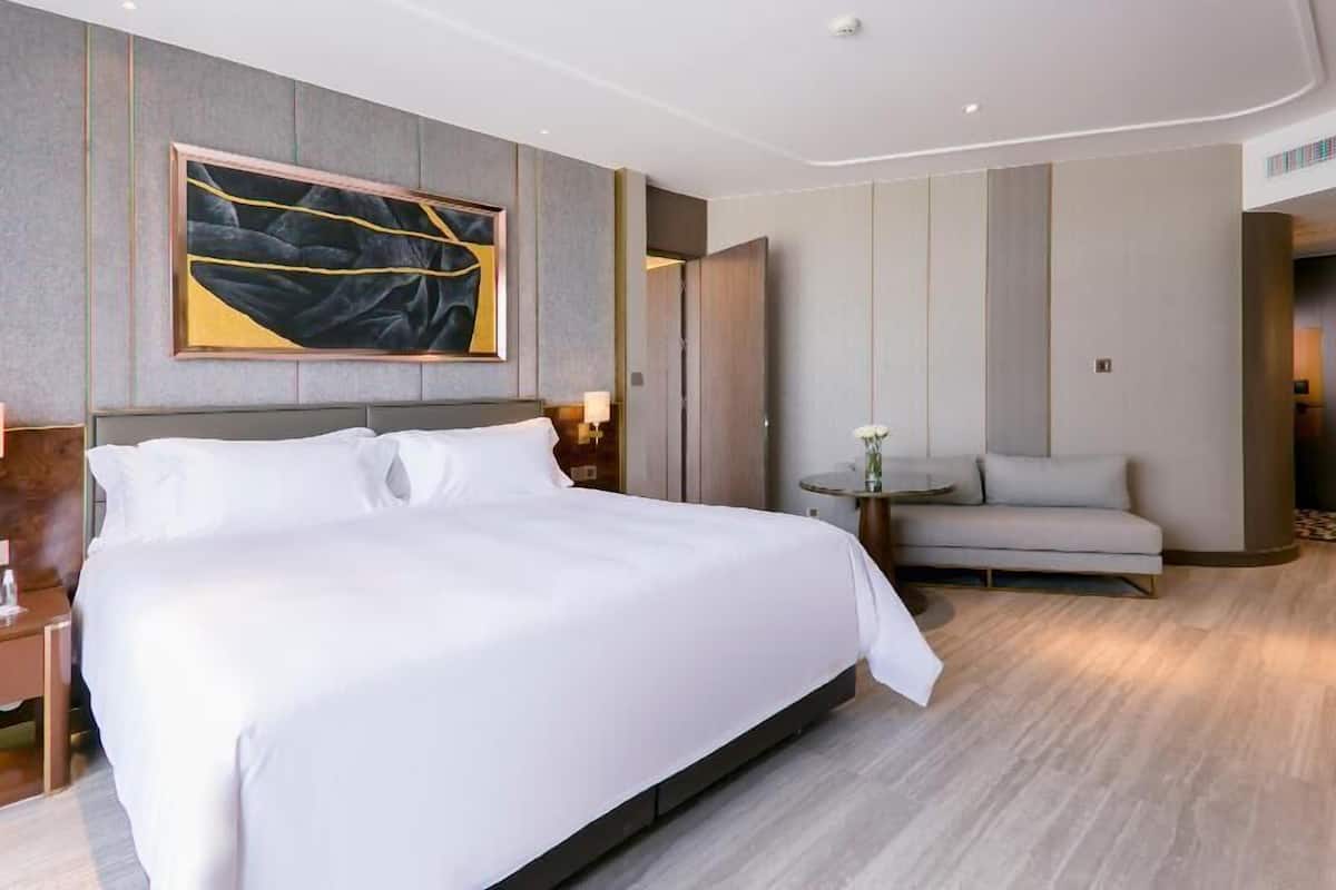 A modern hotel room with a large bed, a sofa, a small table, and a painting on the wall.