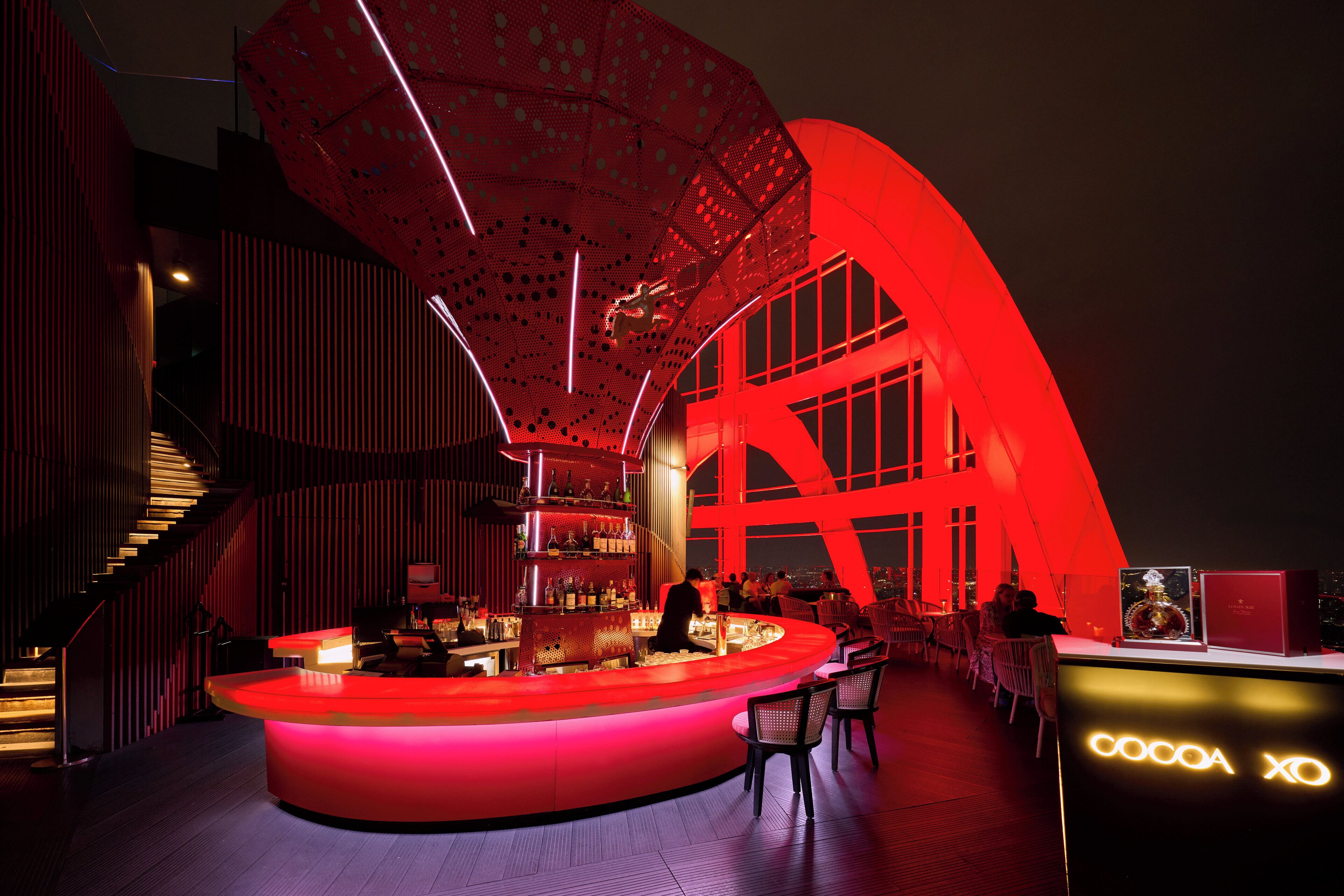 A modern bar area with a circular bar counter, a staircase, and a large red structure in the background.