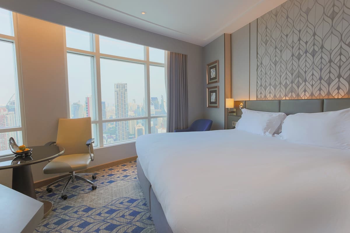 A hotel room with a large bed, a desk with a chair, a window with a city view, and a patterned wall.