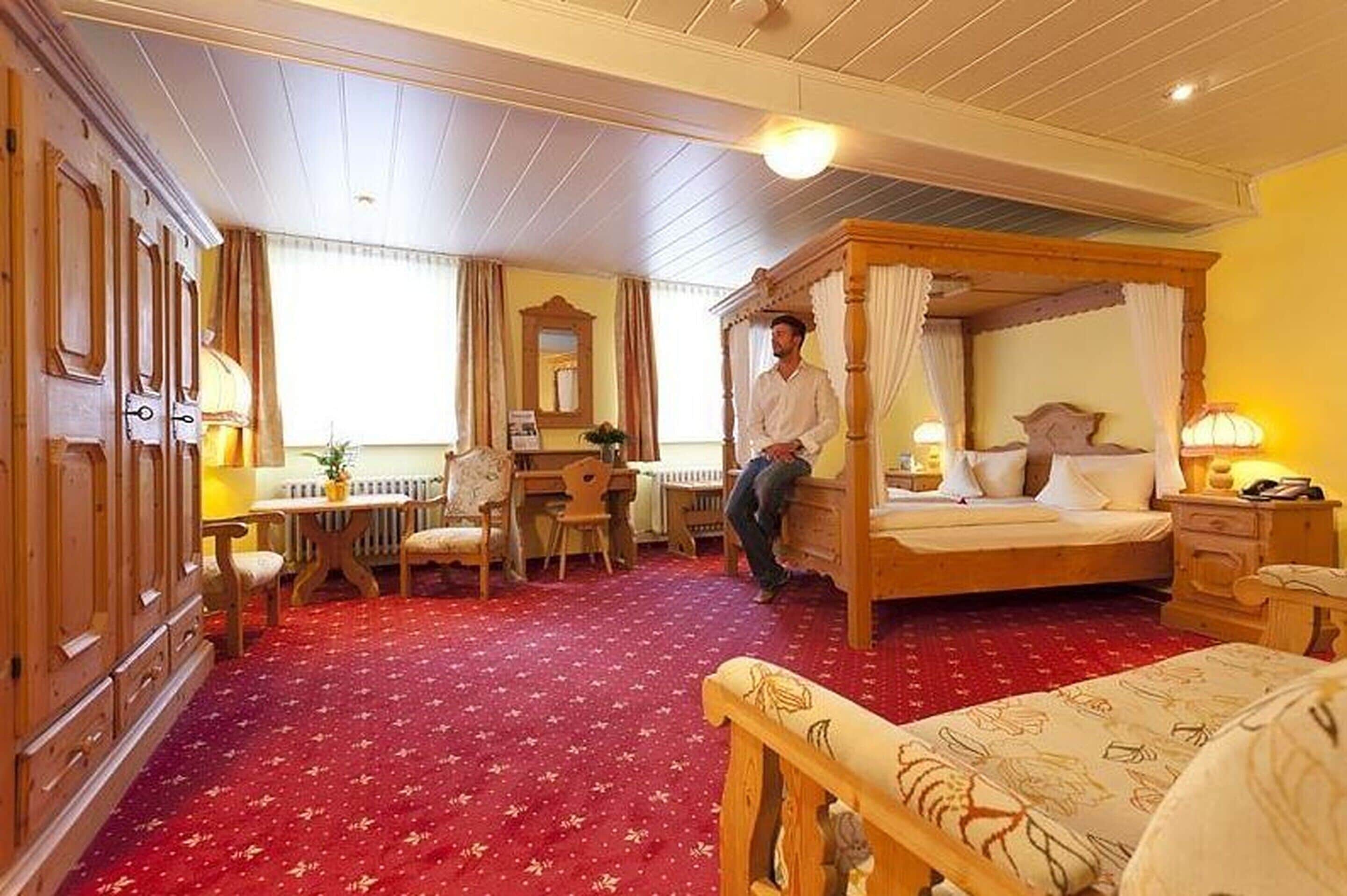 A traditional room with a four-poster bed, a desk with a chair, a radiator, and a red carpet.
