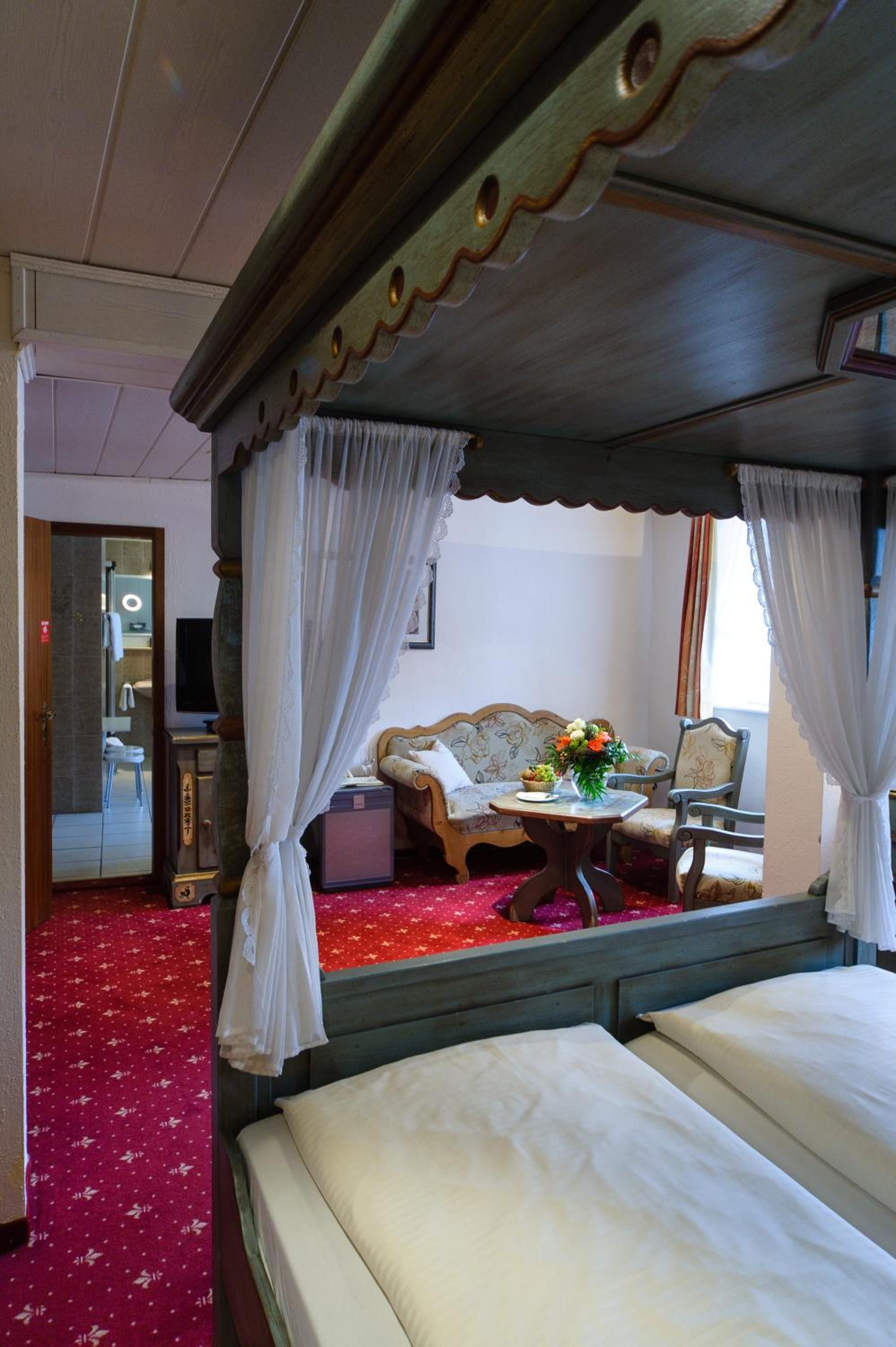 Una cama con dosel y cuatro columnas, una alfombra roja, un sofá y una mesa con flores.