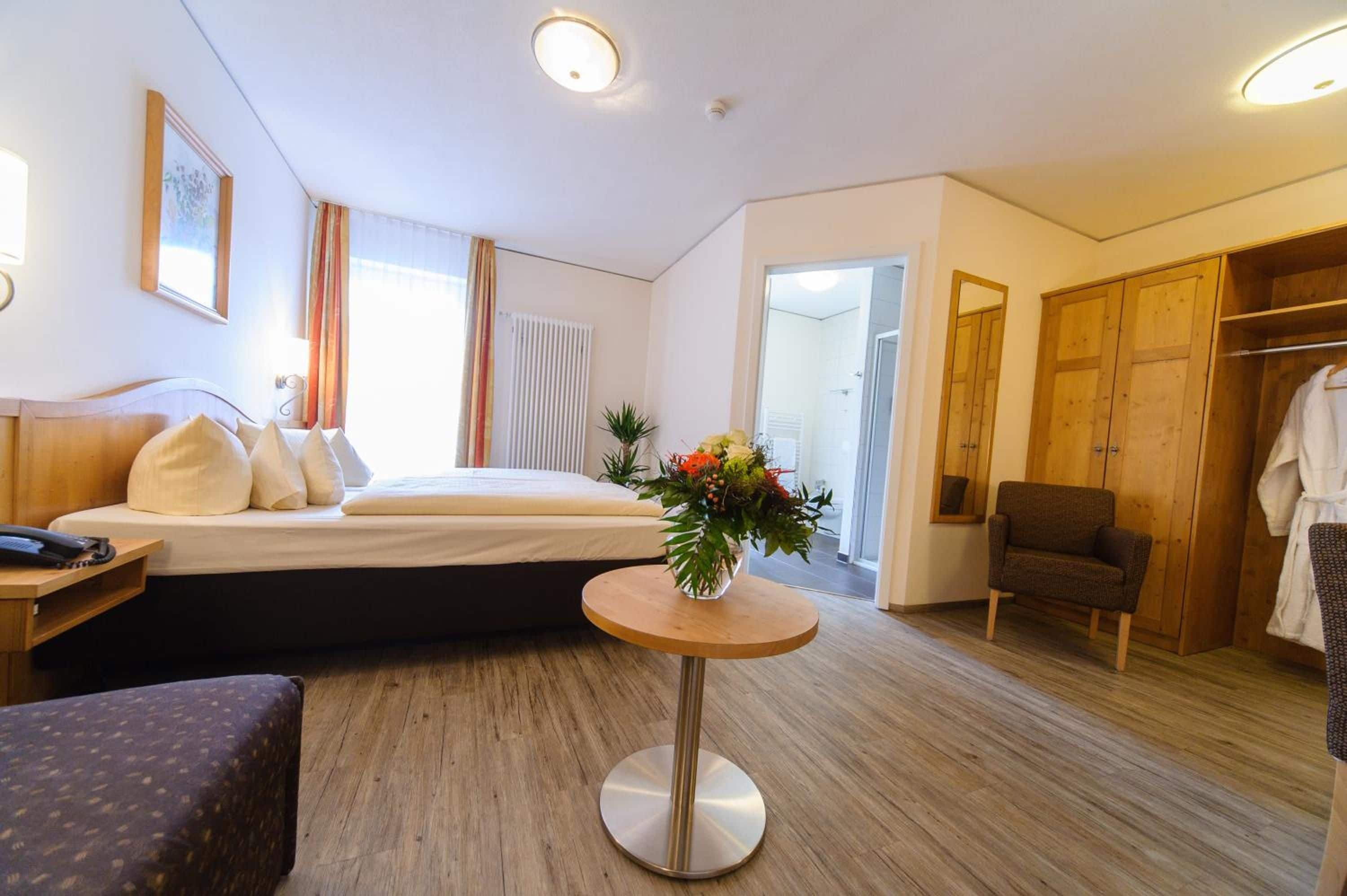 Hotellihuone, jossa on sänky, yöpöytä, tuoli, vaatekaappi ja pieni pyöreä pöytä, jonka päällä on maljakko täynnä kukkia.