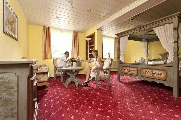 Una habitación con alfombra roja, una cama con dosel, una pequeña mesa con sillas y una estantería.