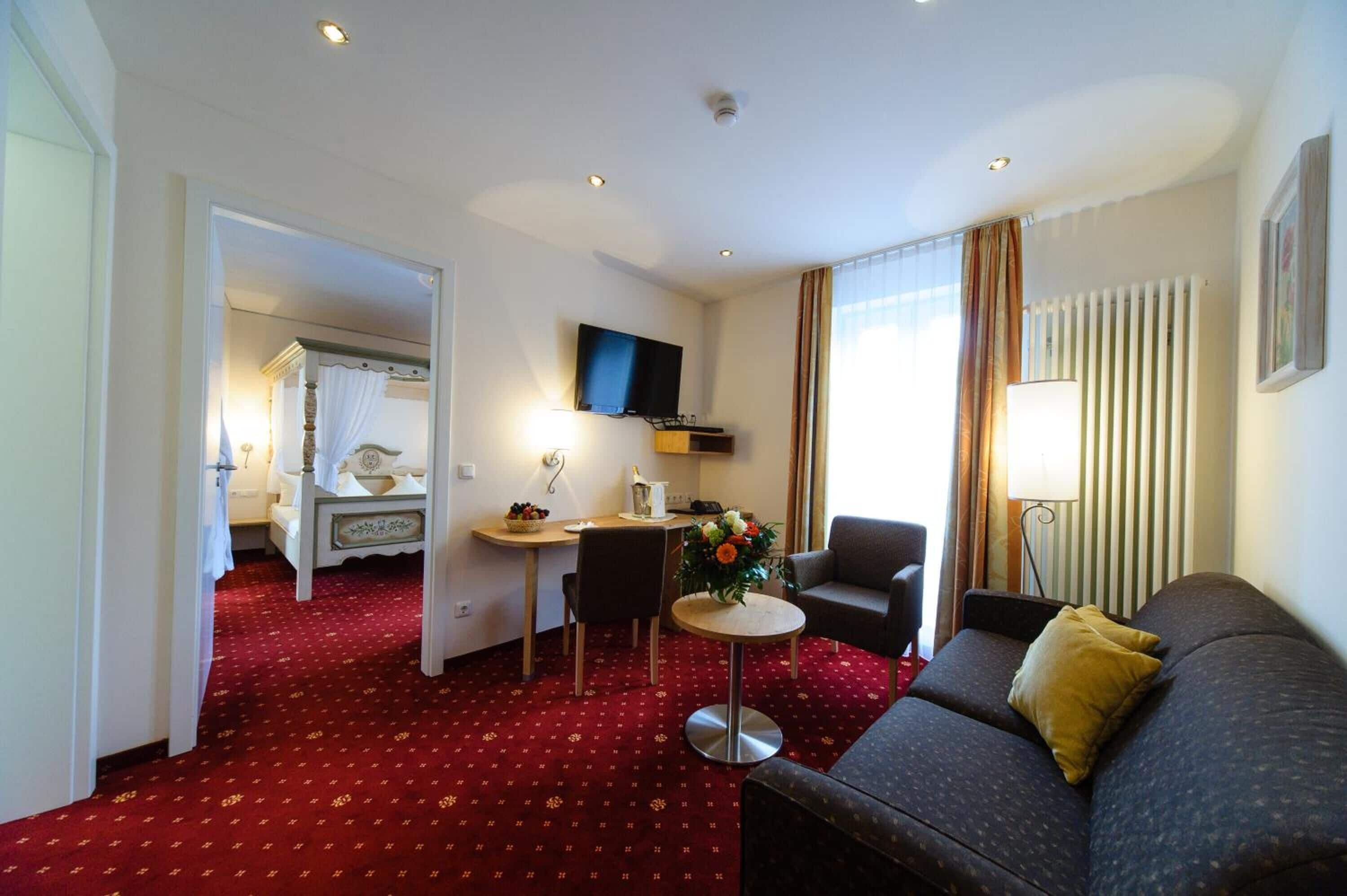 Quarto de hotel com carpete vermelho, sofá, mesinha, televisão, abajur e janela com cortinas.