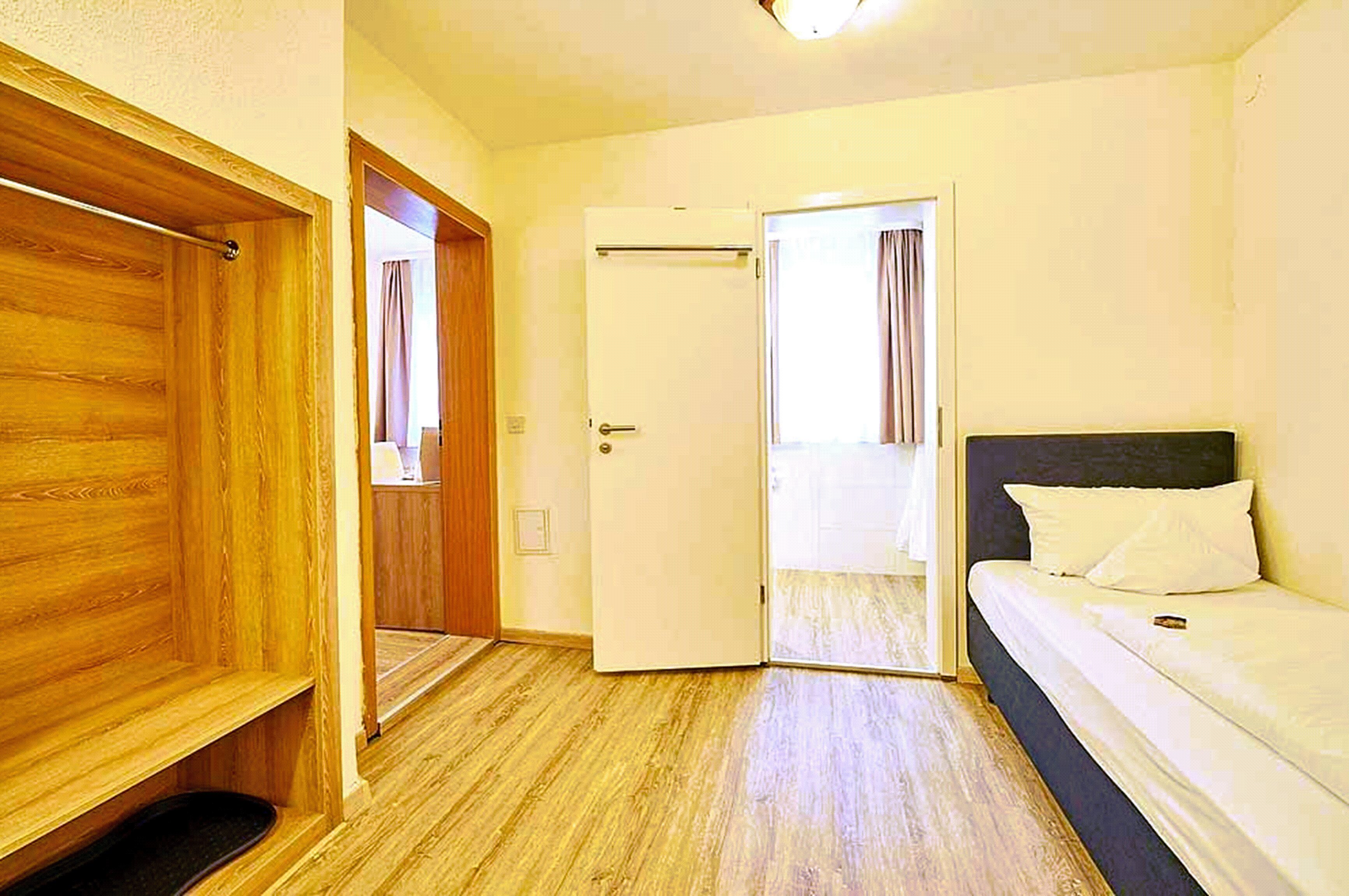 A room with a wooden wardrobe, a bed with white bedding, and a door leading to another room.