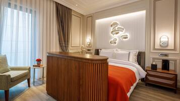 A hotel room with a wooden reception desk, a bed with an orange bedspread, a small side table with a vase of flowers, and a wall-mounted light fixture.