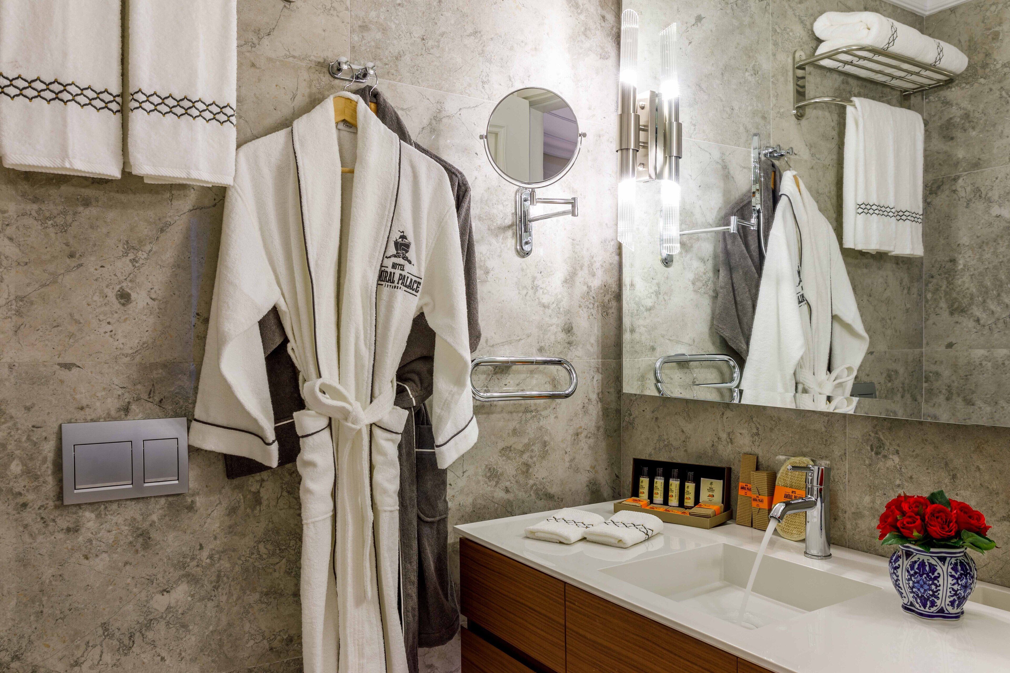 A bathroom with a marble wall, a white robe hanging on a rack, a mirror, a sink with a faucet, and a vase with red flowers.
