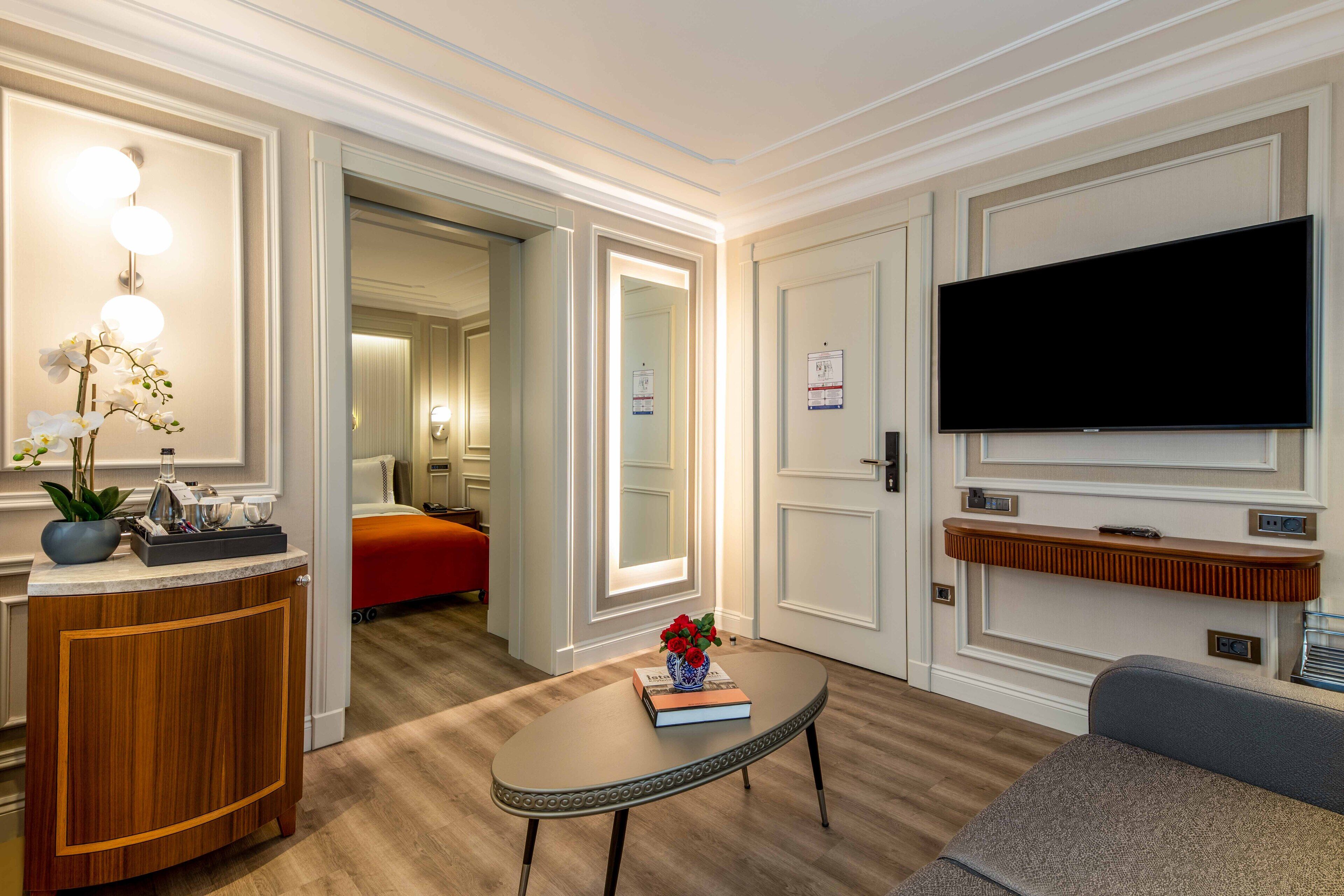 Een moderne hotelkamer met een houten kast, een ronde tafel met een boek en bloemen, een flatscreen televisie en een rode bed.