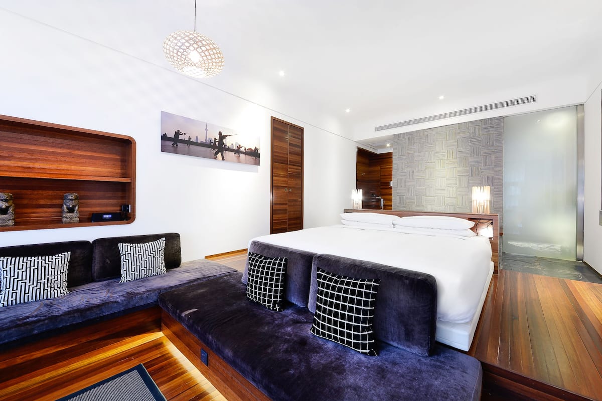 A modern bedroom with a large bed, a wooden bench with cushions, a wall-mounted shelf, and a cityscape picture.