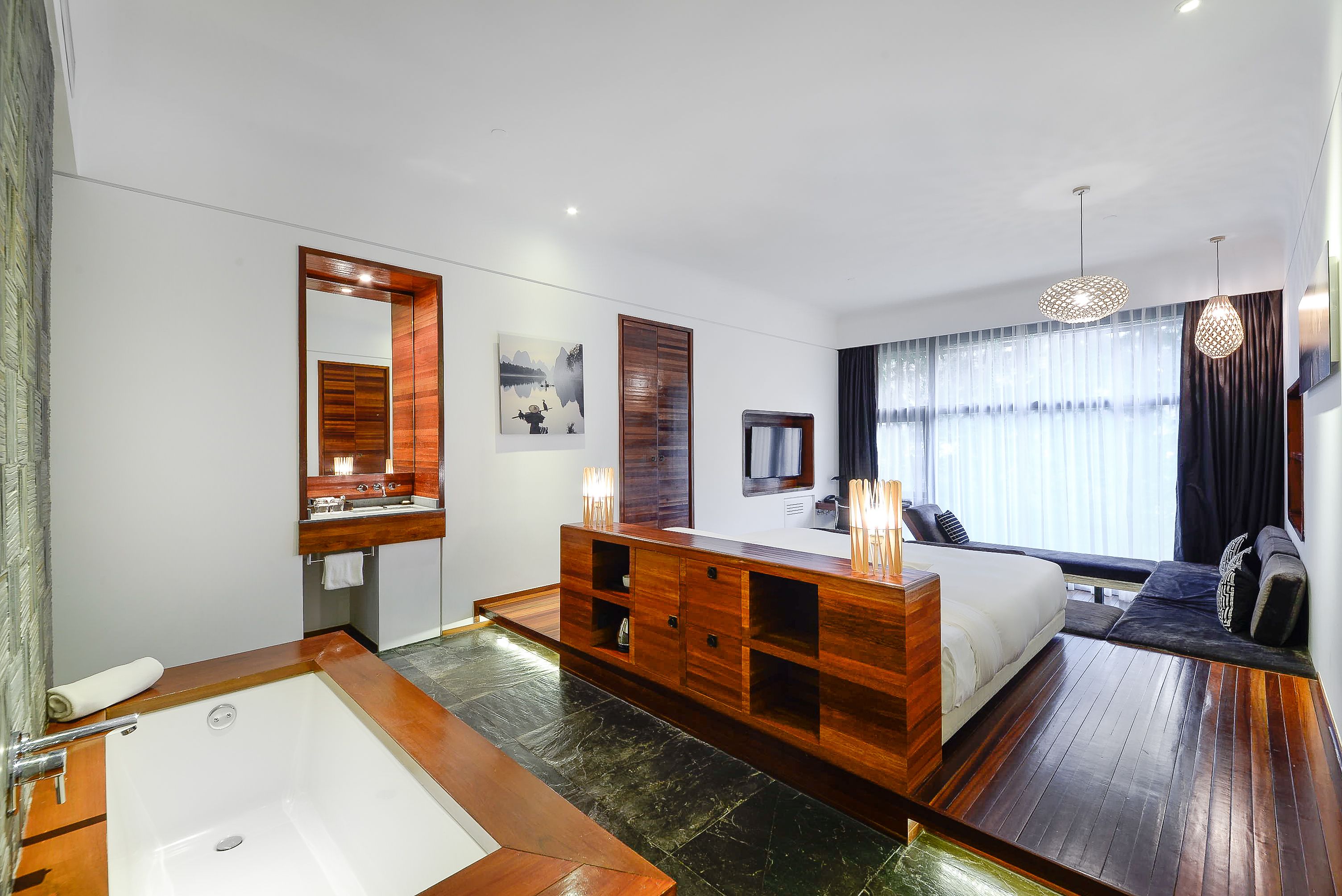 A modern hotel room with a large wooden vanity, a bathtub, a bed with a dark headboard, and a large window with curtains.
