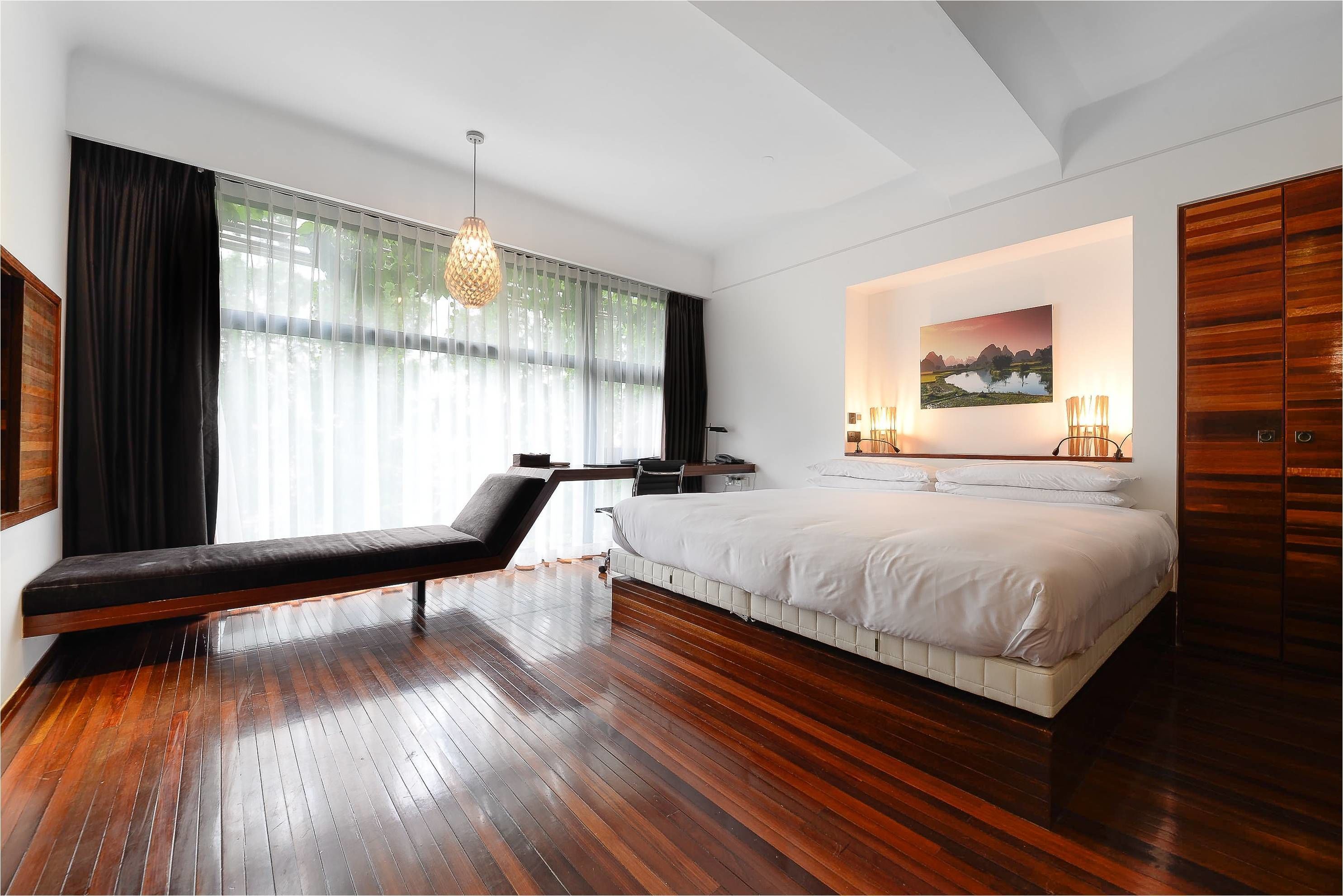 A modern bedroom with a large bed, a bench, a desk, and a large window with curtains.
