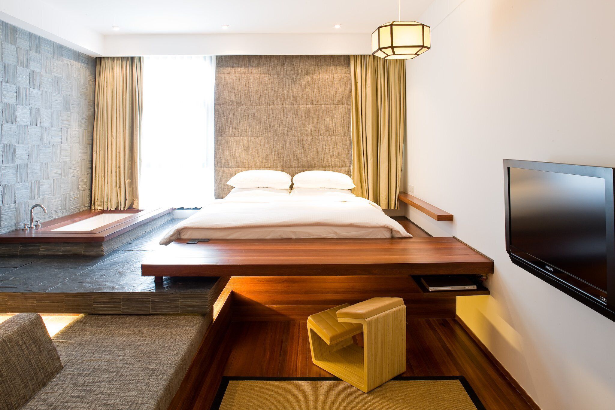 A modern hotel room with a large bed, a built-in TV, a small seating area, and a view of the city.