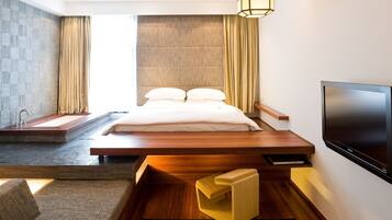 A modern hotel room with a large bed, a built-in TV, a small seating area, and a view of the city.