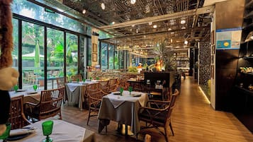 Interior de un restaurante con mesas, sillas, una zona de bar y grandes ventanales con vistas a zonas verdes.
