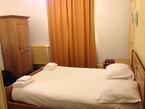 Una cama bien hecha con sábanas y almohadas blancas, un armario de madera, un radiador y una cortina.