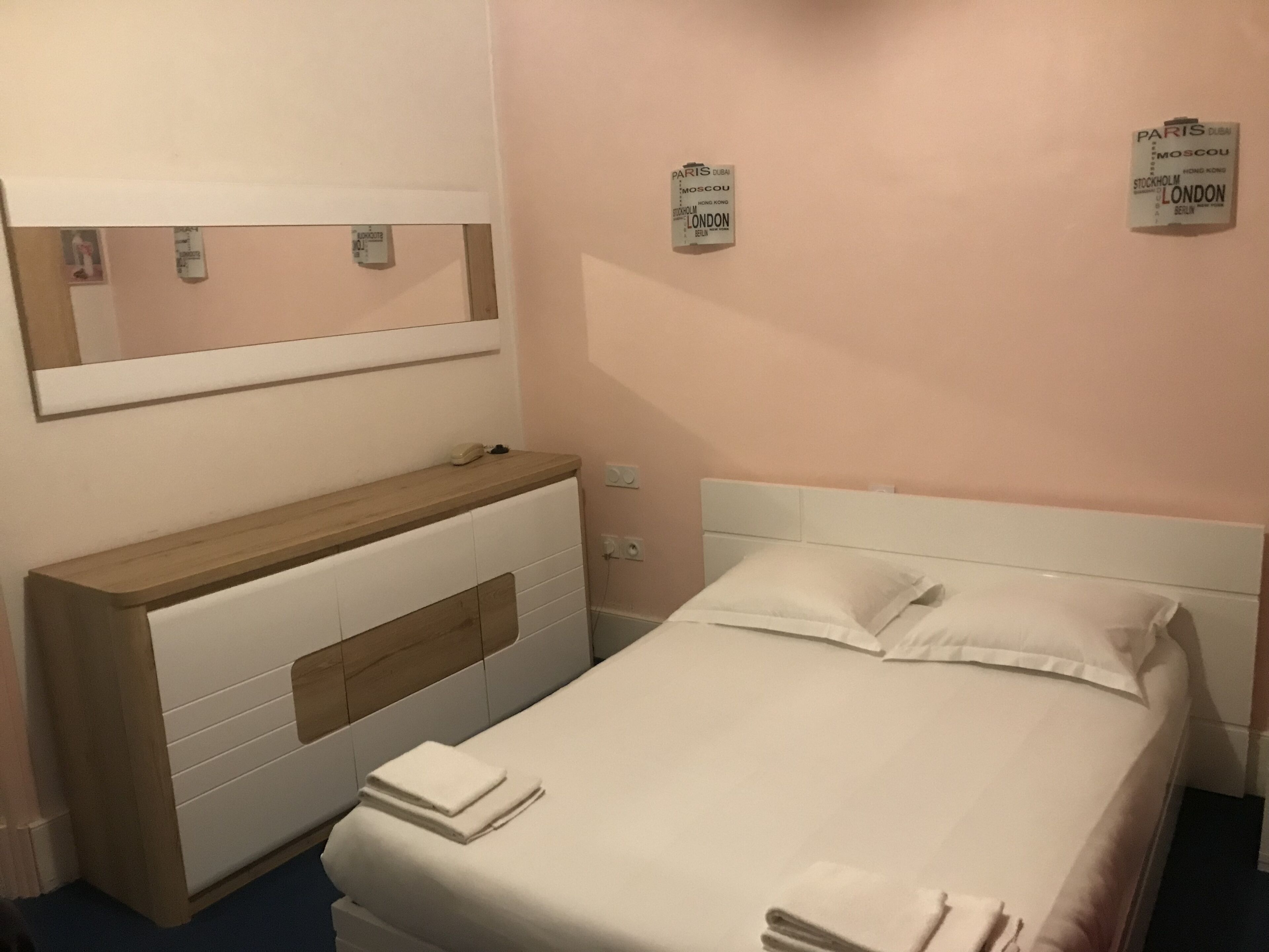 Een hotelkamer met een bed, een houten nachtkastje, een spiegel en een muur met een bord waarop steden staan vermeld.