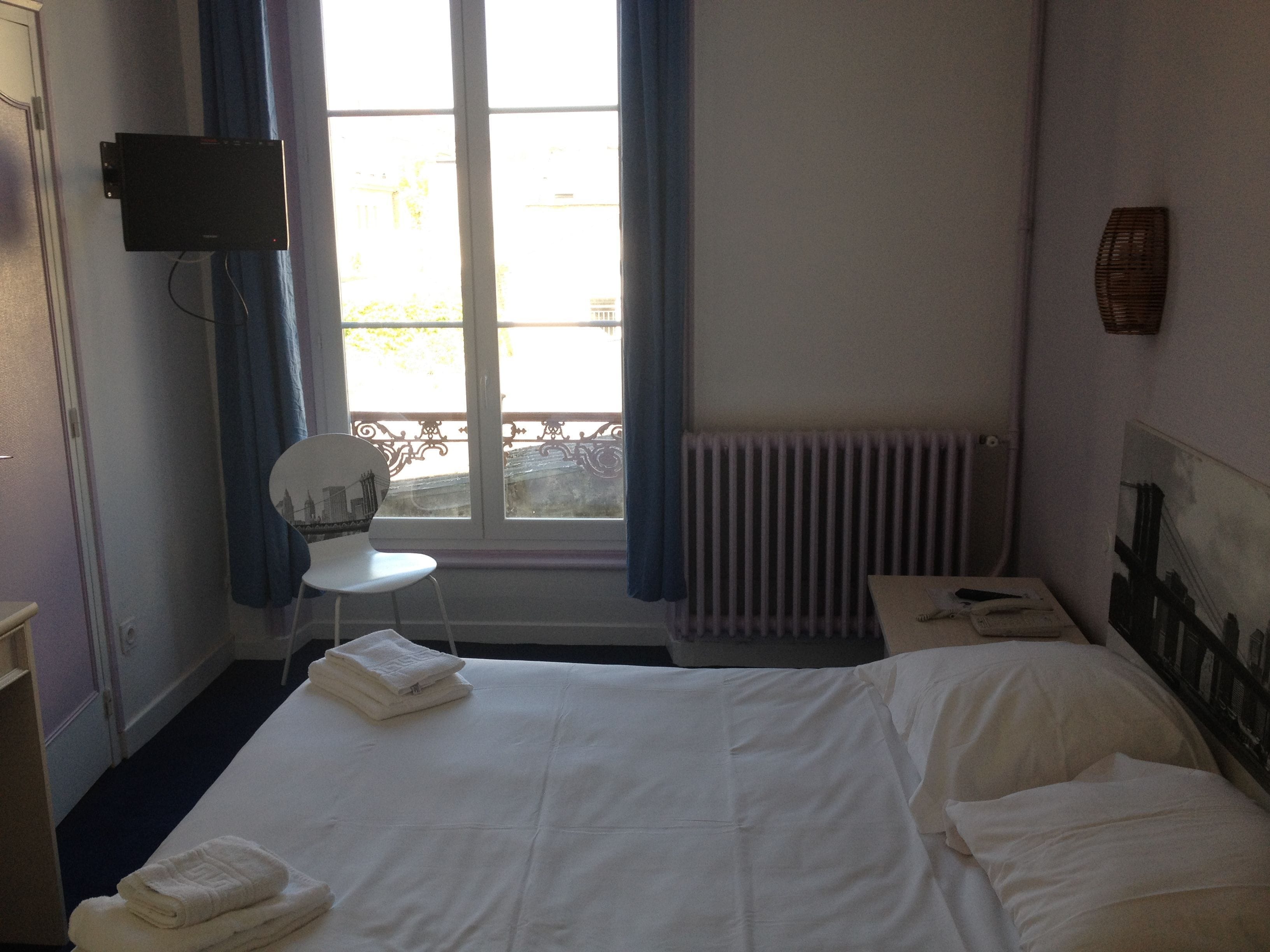 Habitación de hotel con cama, silla, una mesita, televisor montado en la pared y ventana con cortinas.