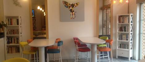 A cozy cafe with a white table, colorful chairs, a bookshelf, and a decorative wall piece.