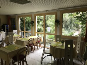 Un comedor con mesas y sillas, ventanas amplias con plantas y una cafetera.