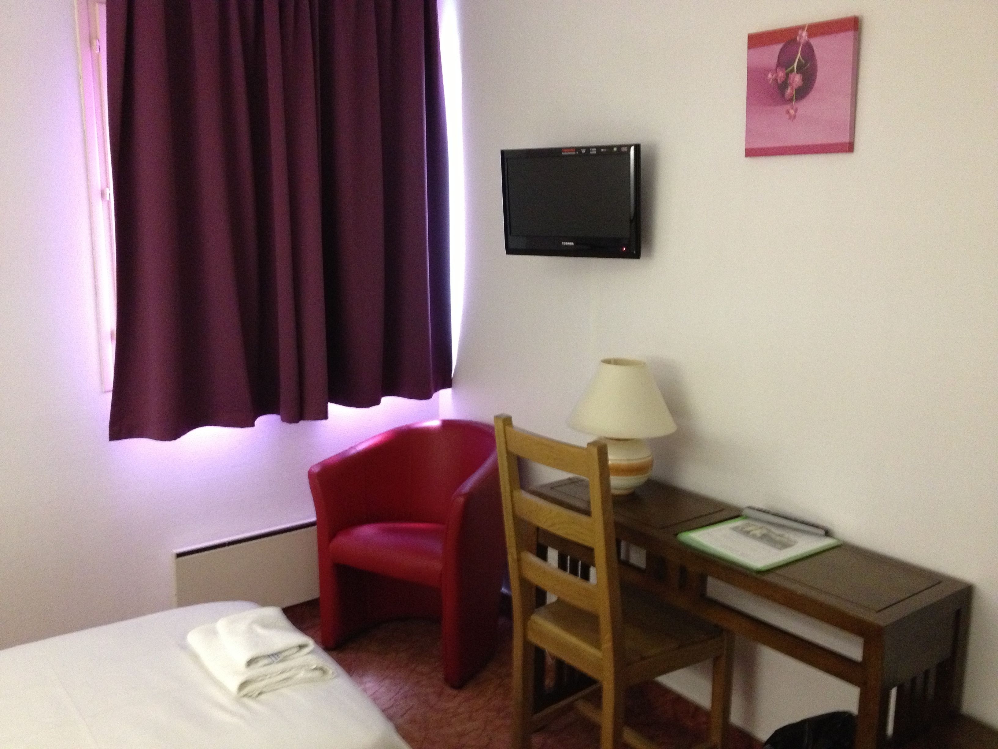 Una camera d'albergo con un letto, una scrivania con lampada, una sedia rossa, una televisione e una finestra con tende.