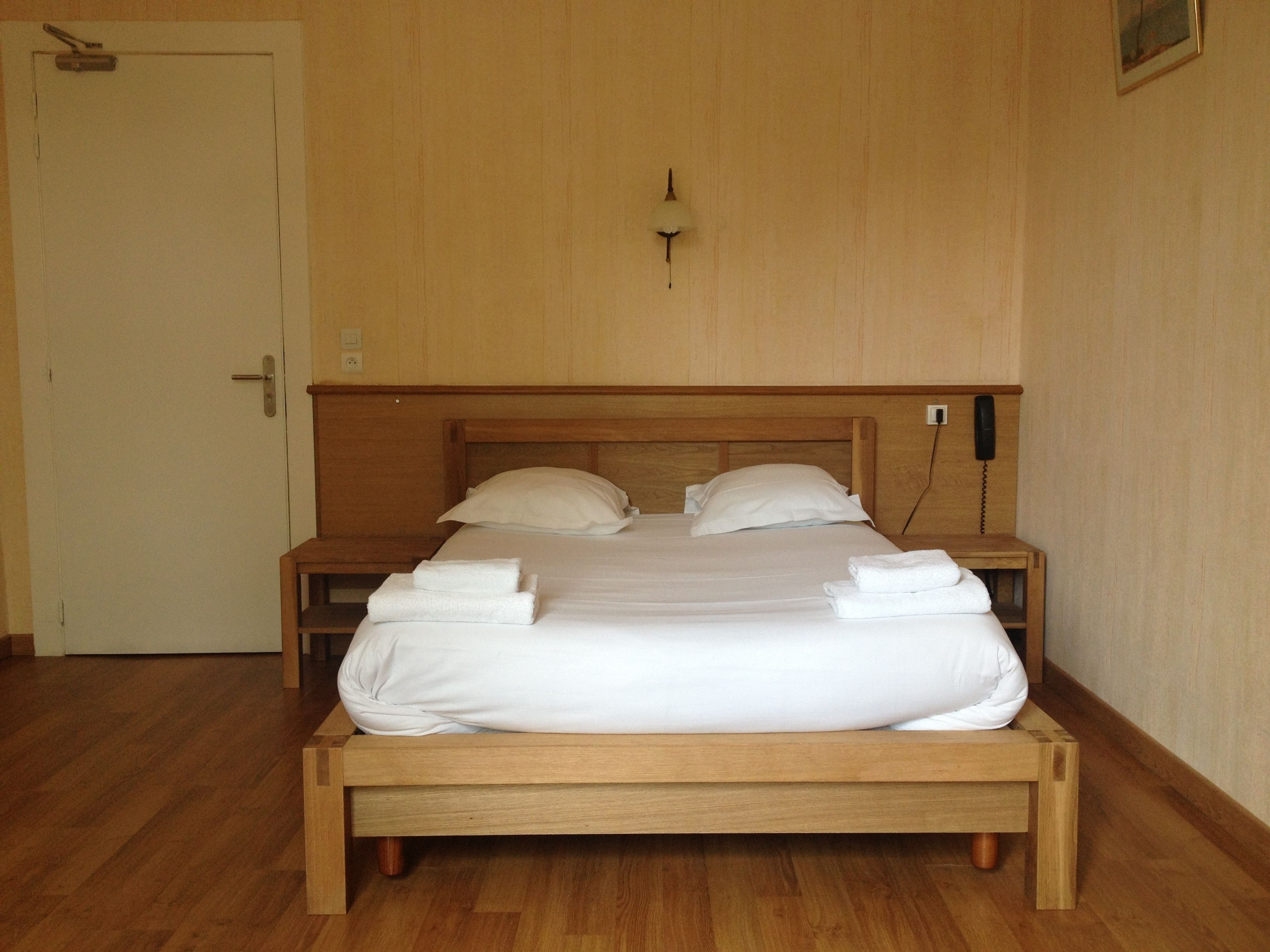 A wooden bed with white linens and towels, a bedside table, a wall-mounted phone, and a door.