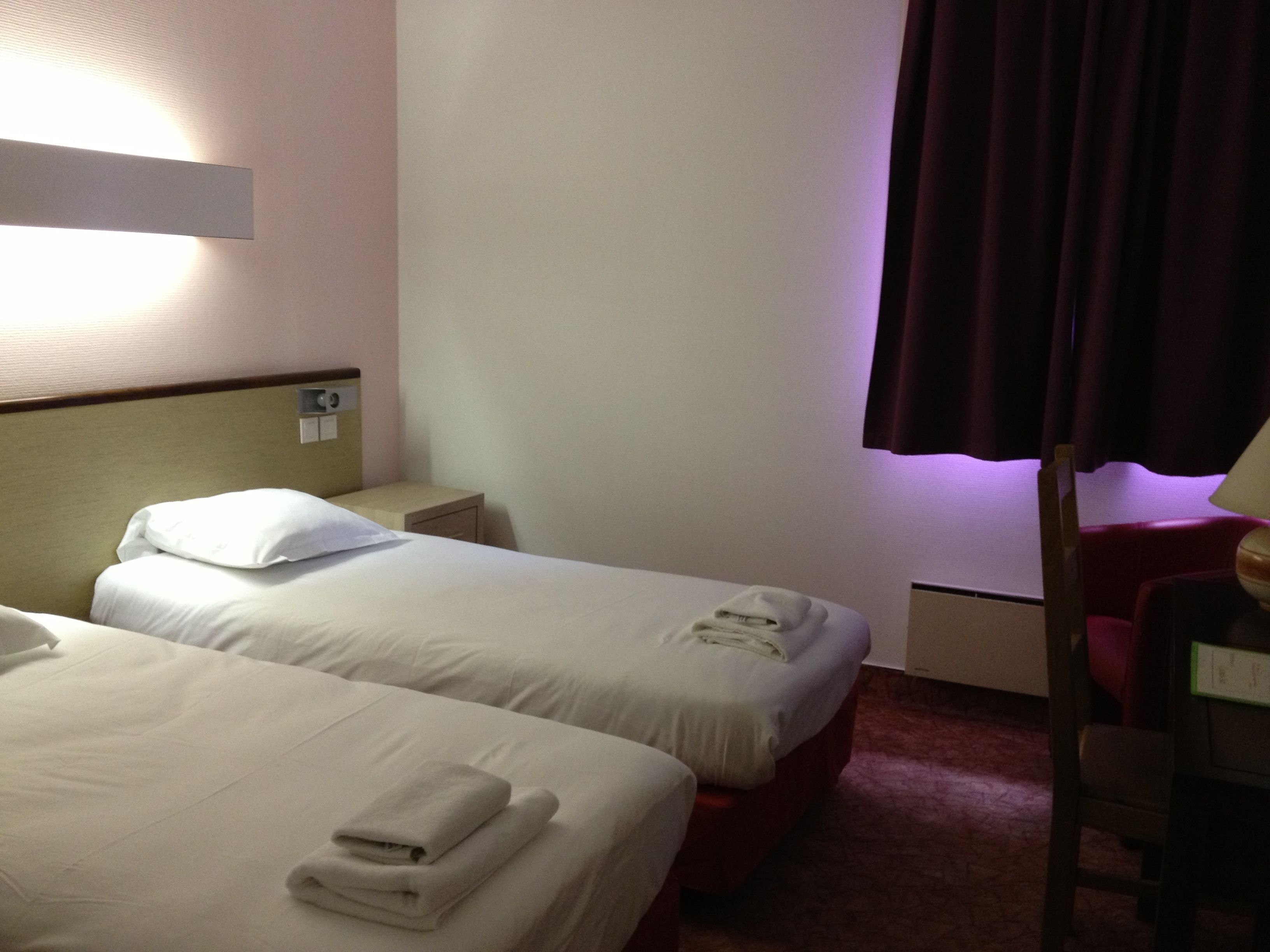 A hotel room with two beds, a nightstand, a chair, and a window with curtains.