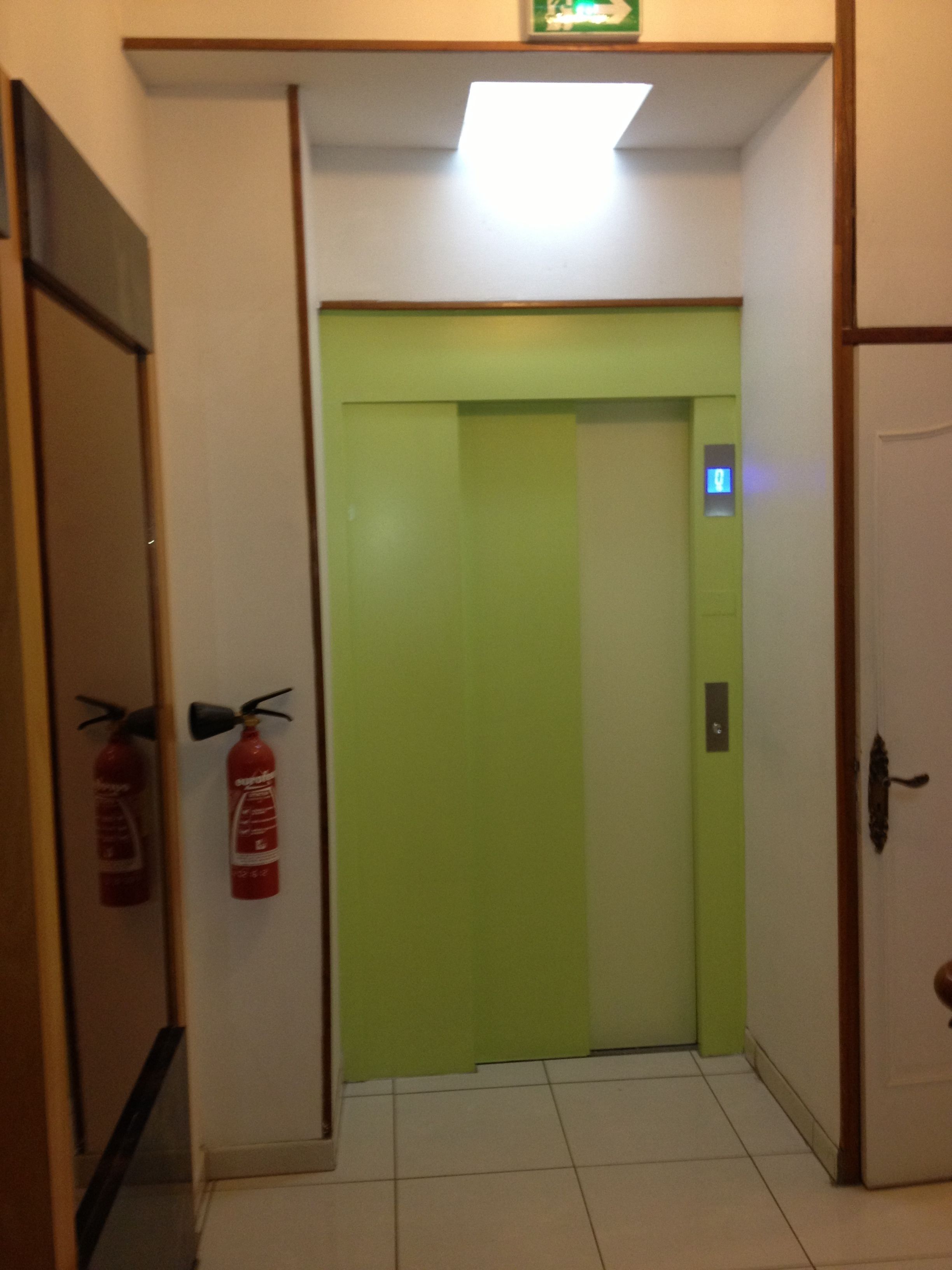 Une porte d’ascenseur verte, avec un extincteur fixé au mur sur le côté gauche.