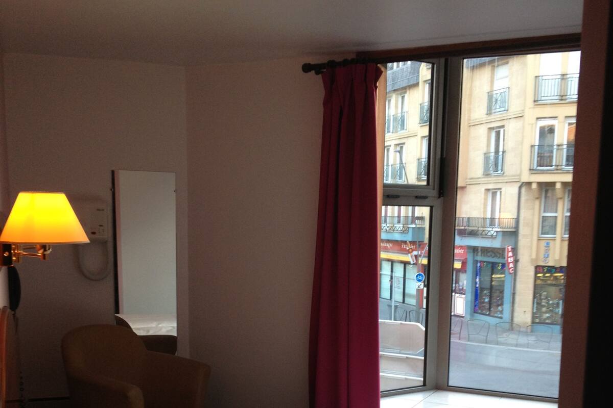 Une chambre d’hôtel avec un lit, une chaise, une lampe, une fenêtre donnant sur des immeubles et un rideau.