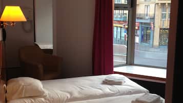 Une chambre d’hôtel avec un lit, une chaise, une lampe, une fenêtre donnant sur des immeubles et un rideau.