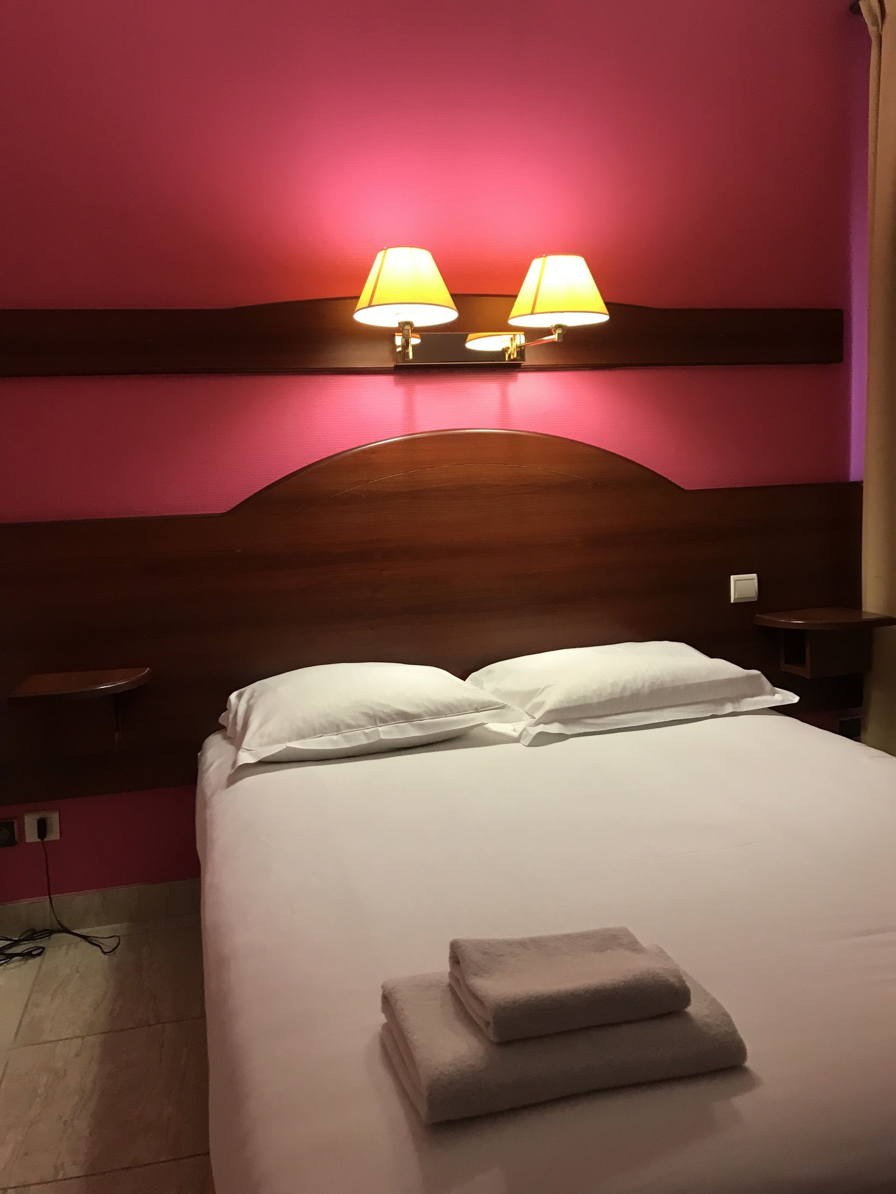 A hotel room with a bed, two bedside lamps, a wooden headboard, and a nightstand.