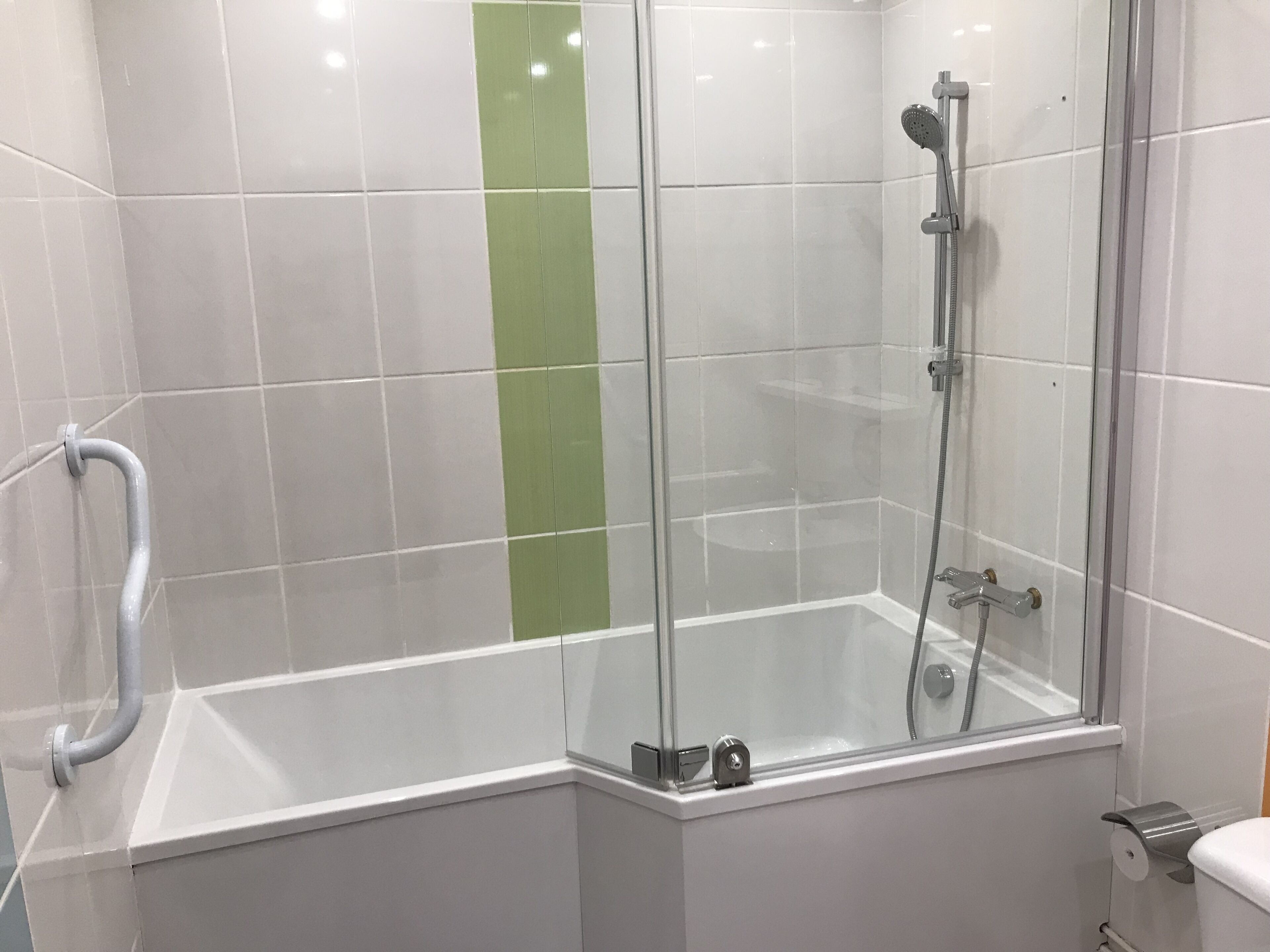 Un área de ducha con paredes de vidrio, una bañera blanca, una franja vertical verde en la pared y un pasamanos.