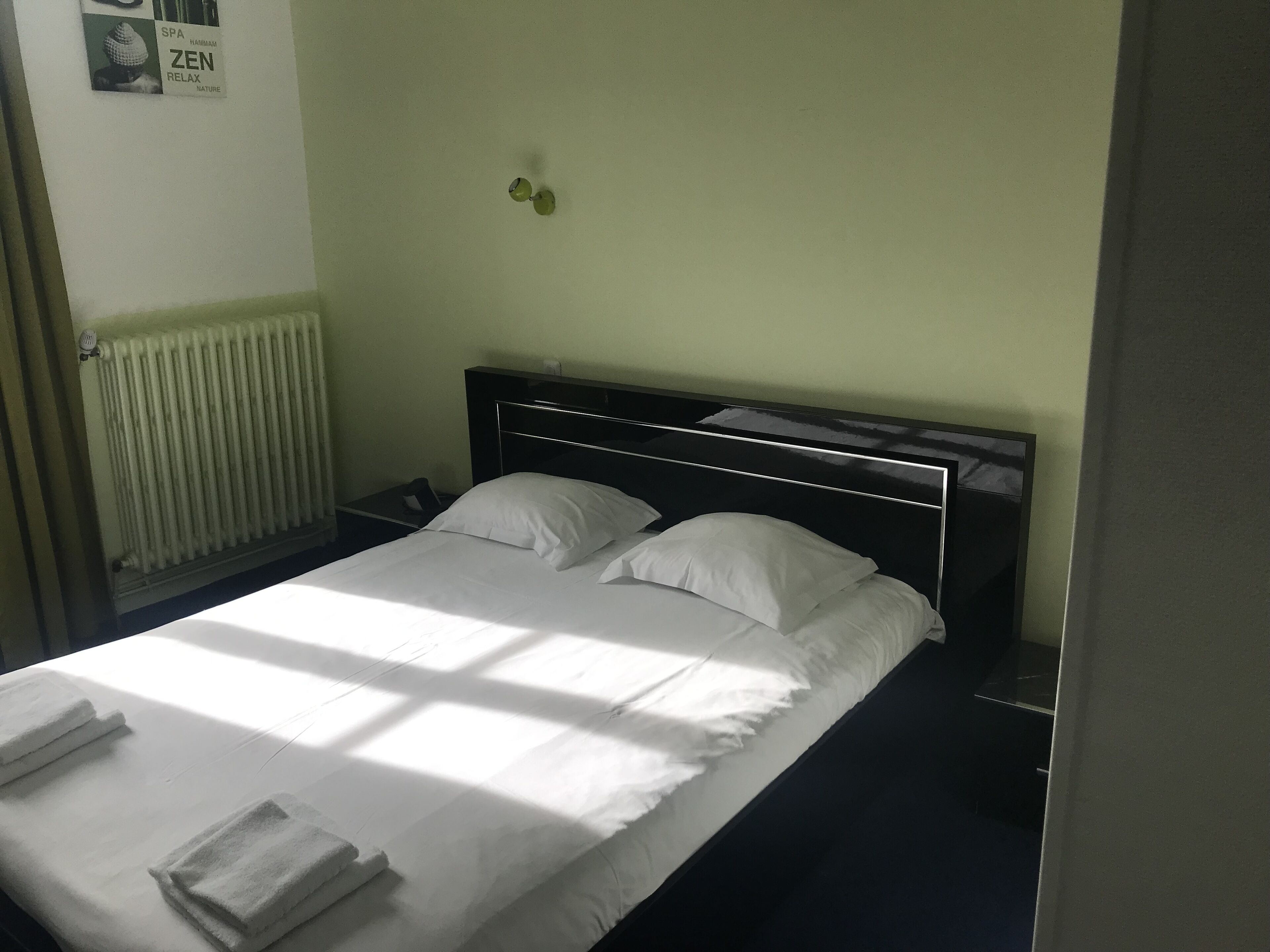 Una cama bien hecha con sábanas y almohadas blancas, un radiador y una pared con un cartel.
