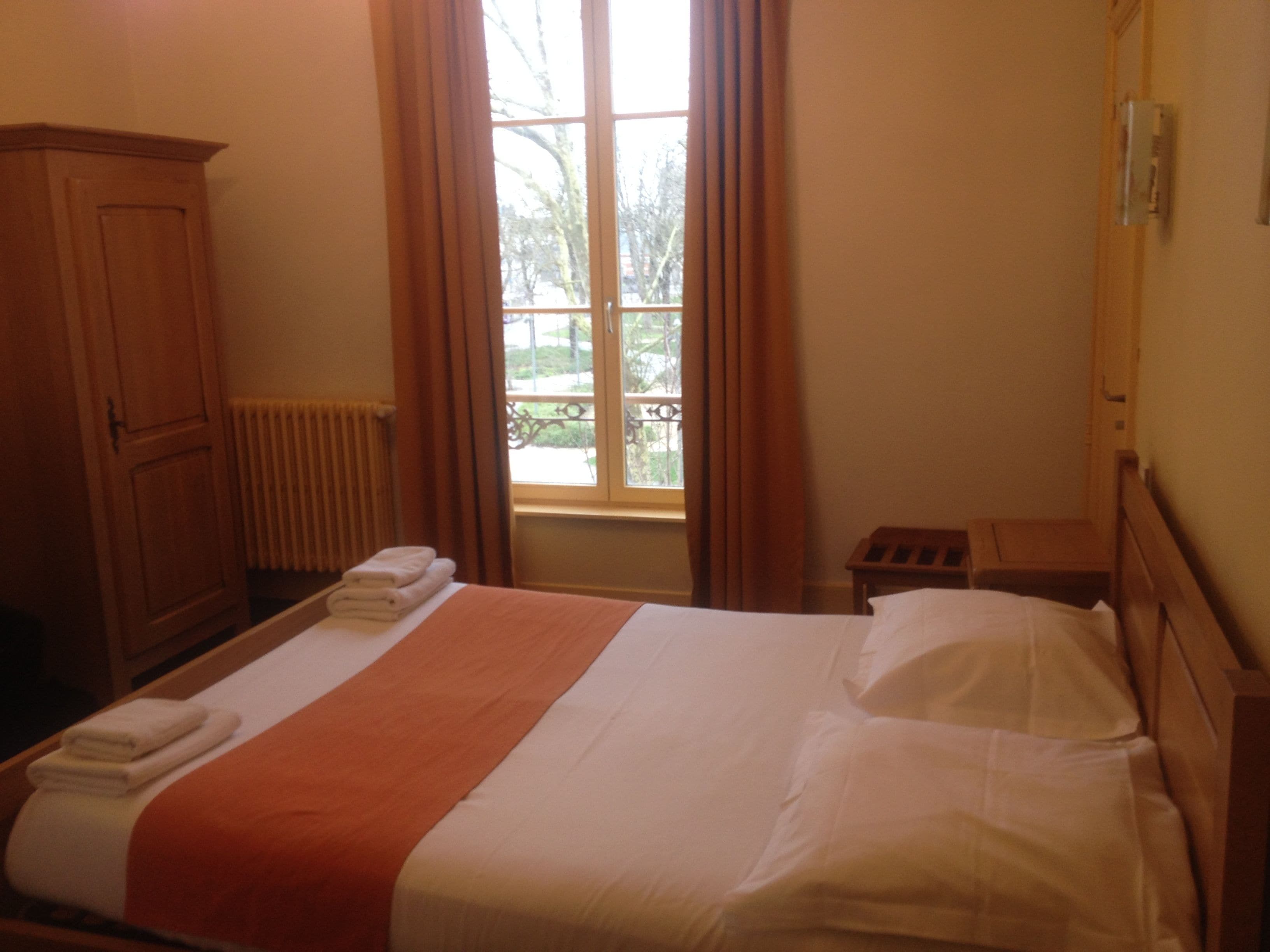 A hotel room with a bed, a wooden wardrobe, a window with curtains, and a small bedside table.