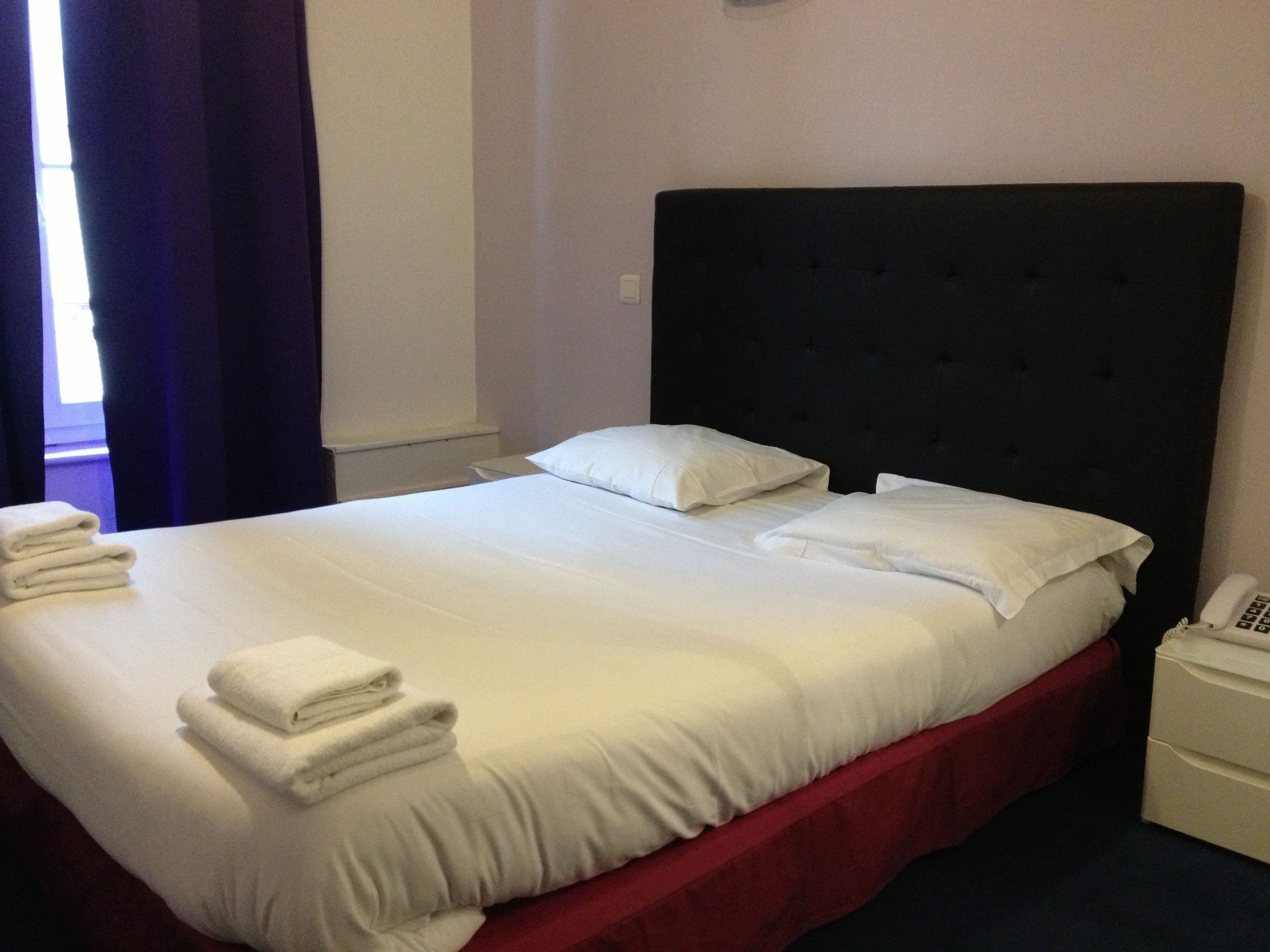A neatly made bed with white linens and pillows, a dark headboard, and a small bedside table with a telephone.