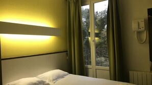A hotel room with a bed, a bedside lamp, a window with curtains, and a telephone.
