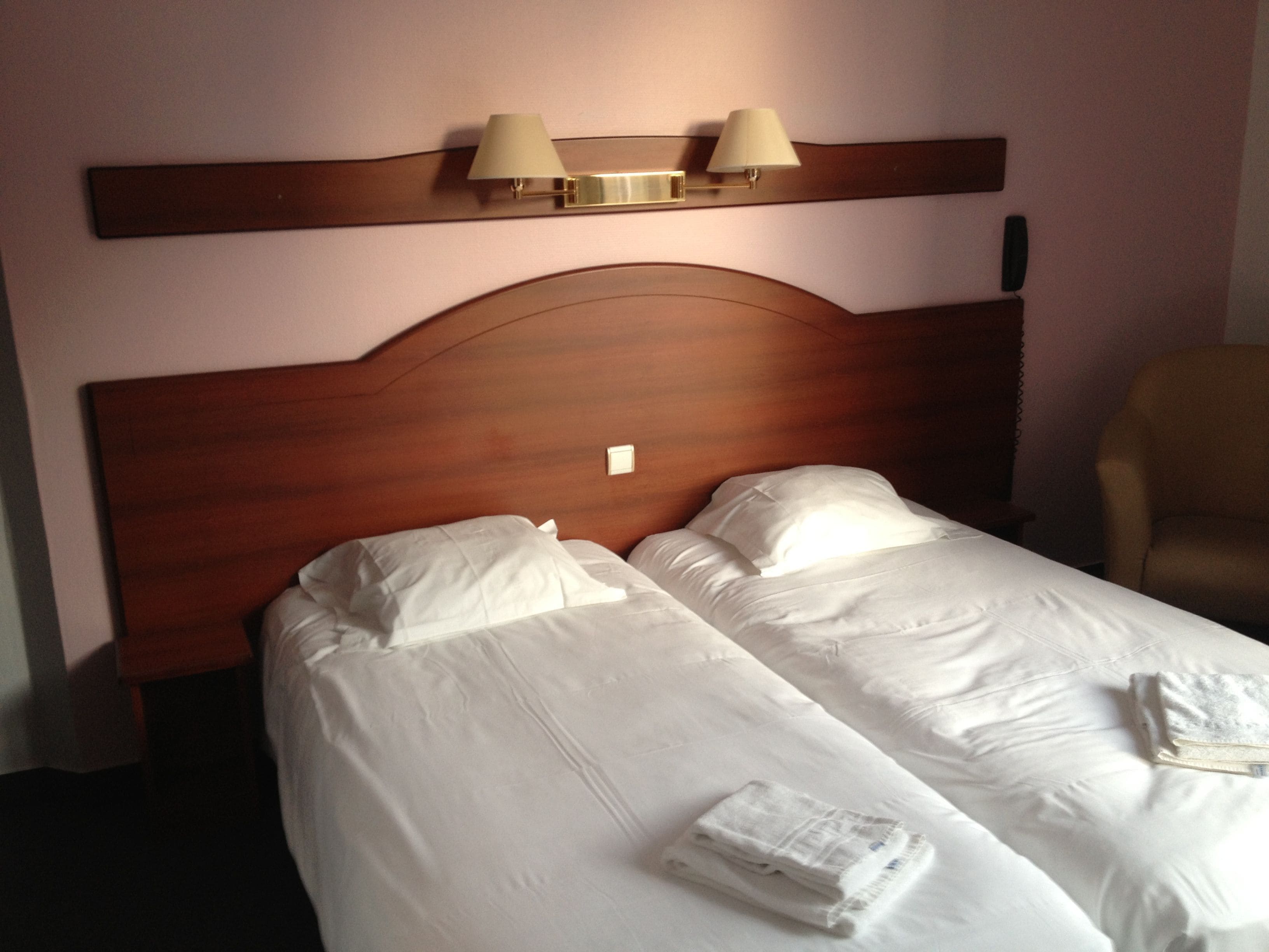 Une chambre d’hôtel avec une tête de lit en bois, deux lits avec du linge de lit blanc et deux lampes fixées au mur.