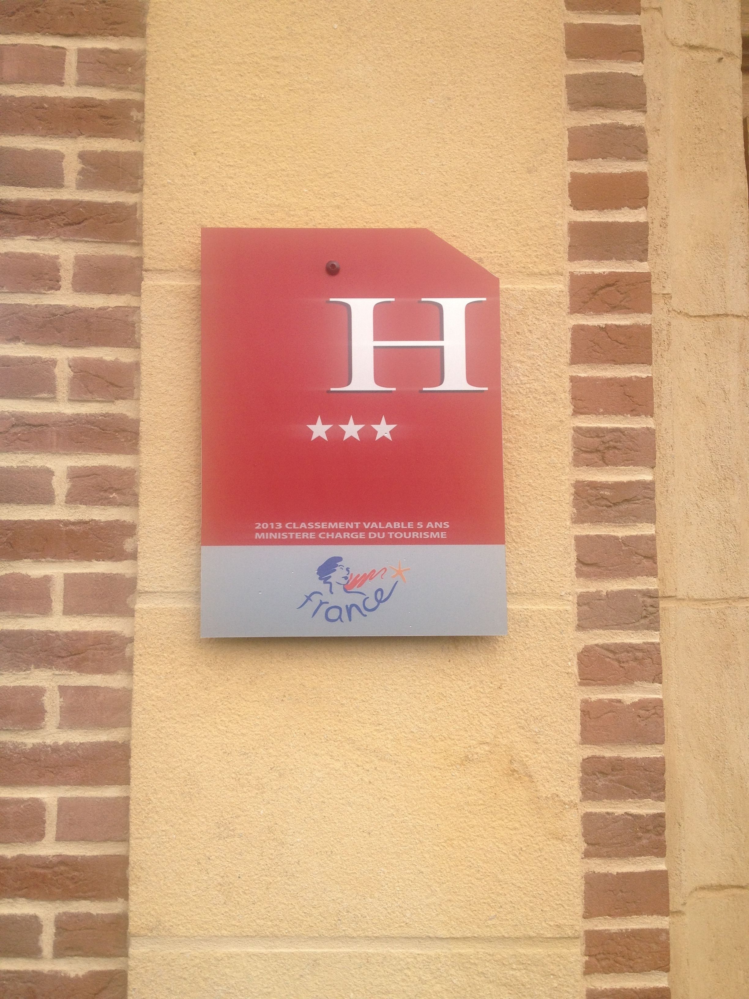 En röd plakett med ett stort ”H” och tre stjärnor, monterad på en tegelvägg.