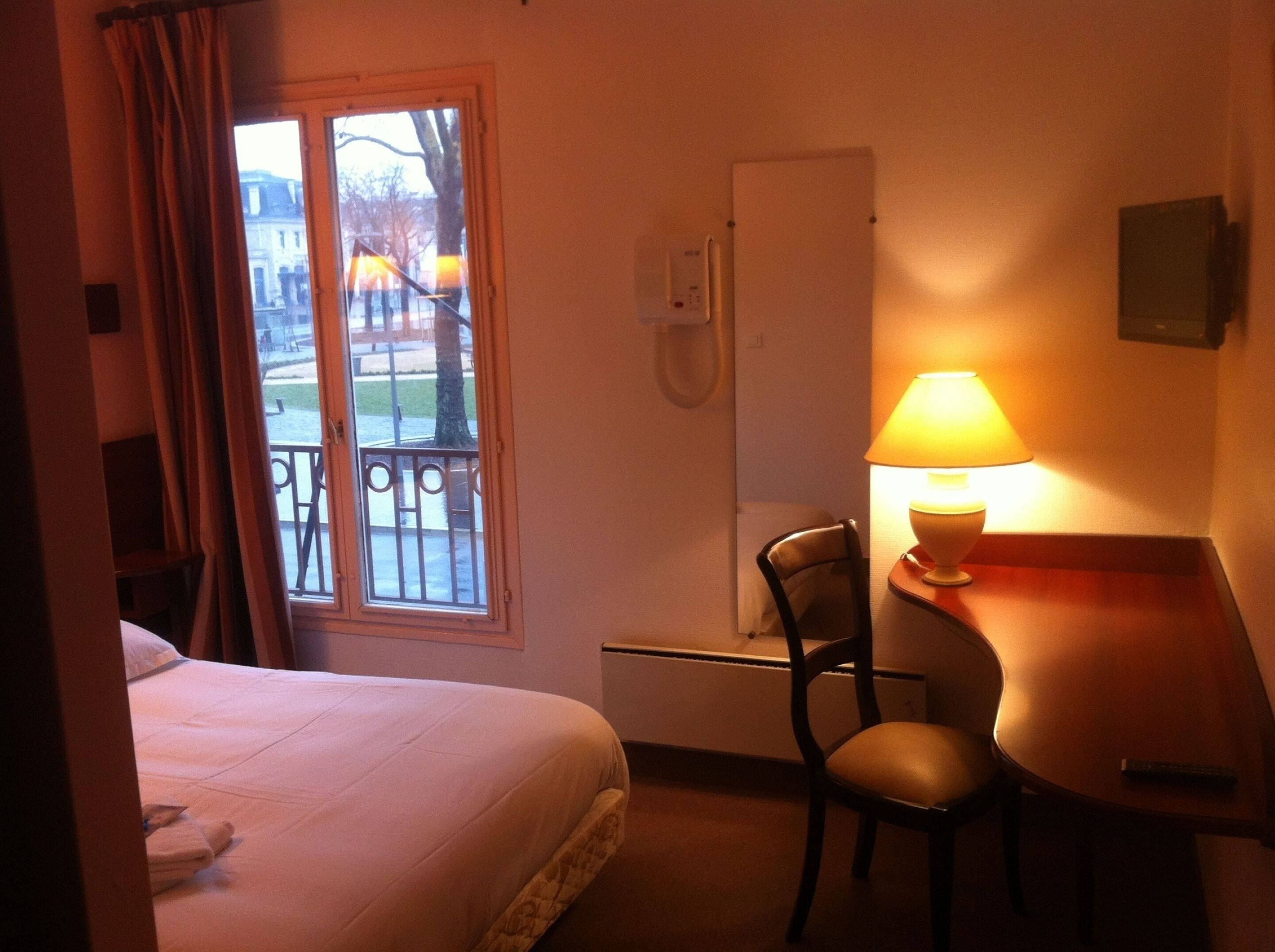 Una habitación de hotel con cama, escritorio, silla, lámpara y ventana con vistas a edificios y árboles.