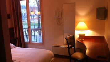 Una habitación de hotel con cama, escritorio, silla, lámpara y ventana con vistas a edificios y árboles.