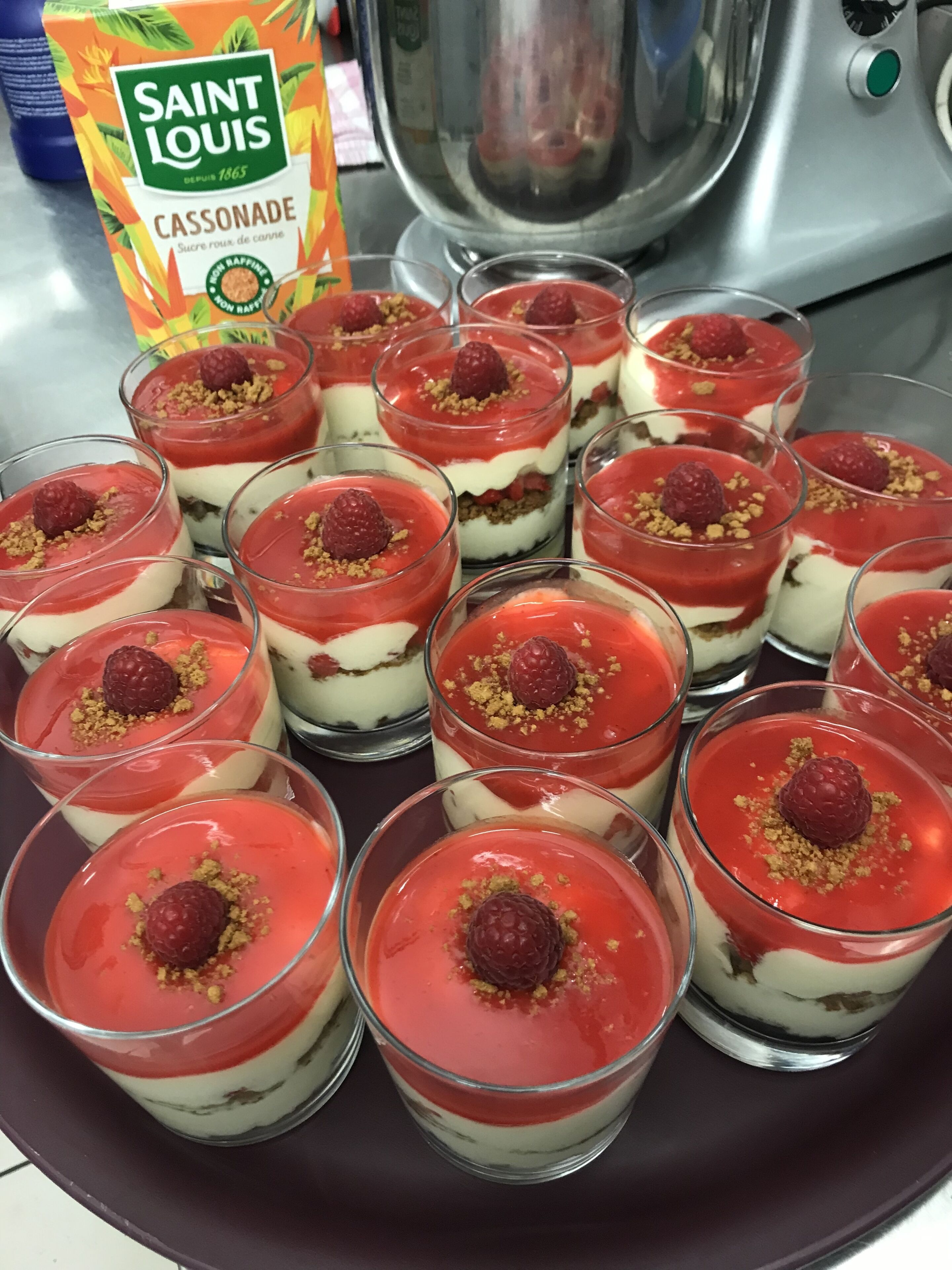 A tray of dessert cups with a raspberry topping and crumbled biscuit, a bag of Saint Louis Cassonade, and a kitchen appliance in the background.
